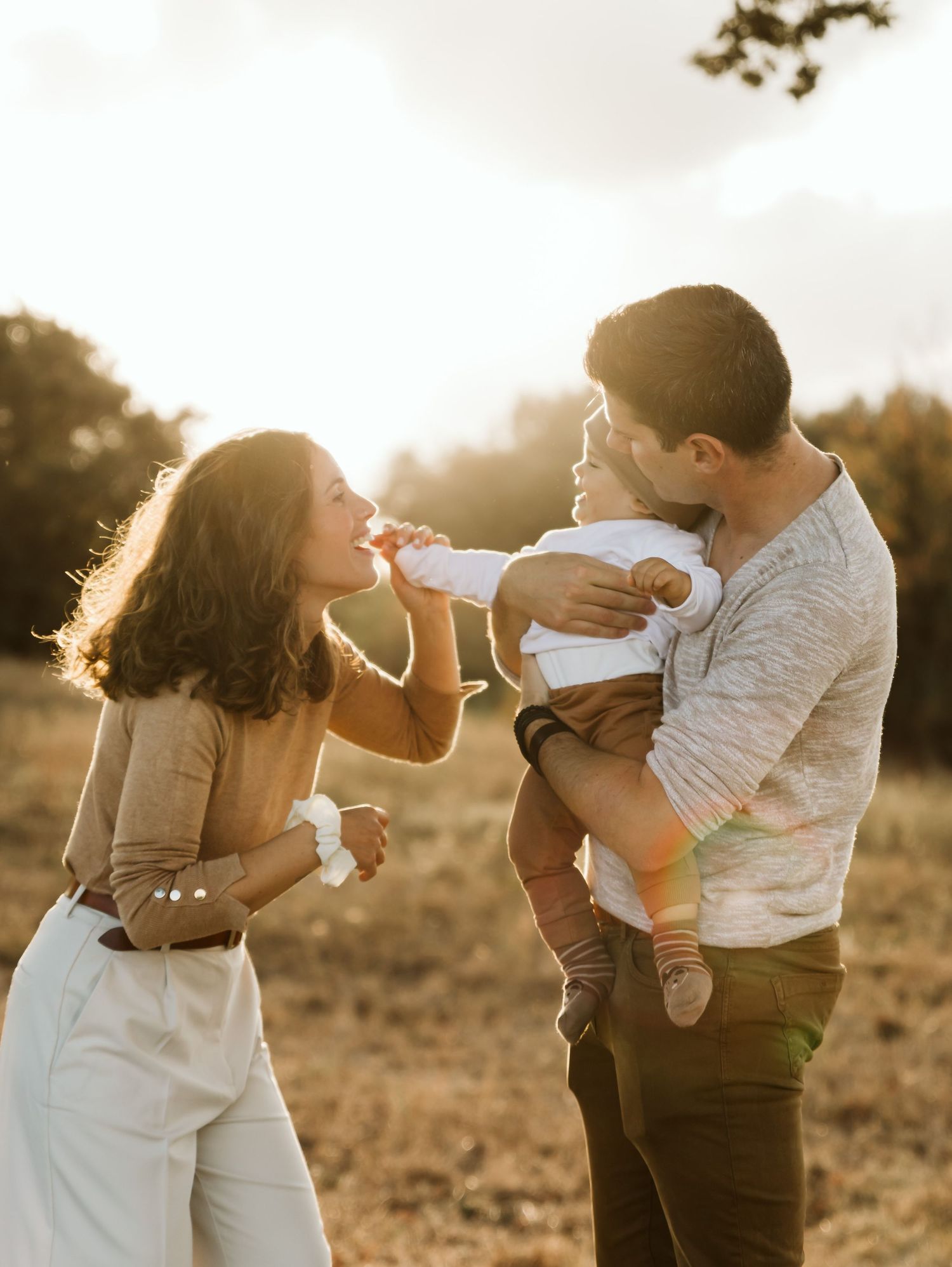 Father holding baby who reaches toward mother in a warm golden sunset field.