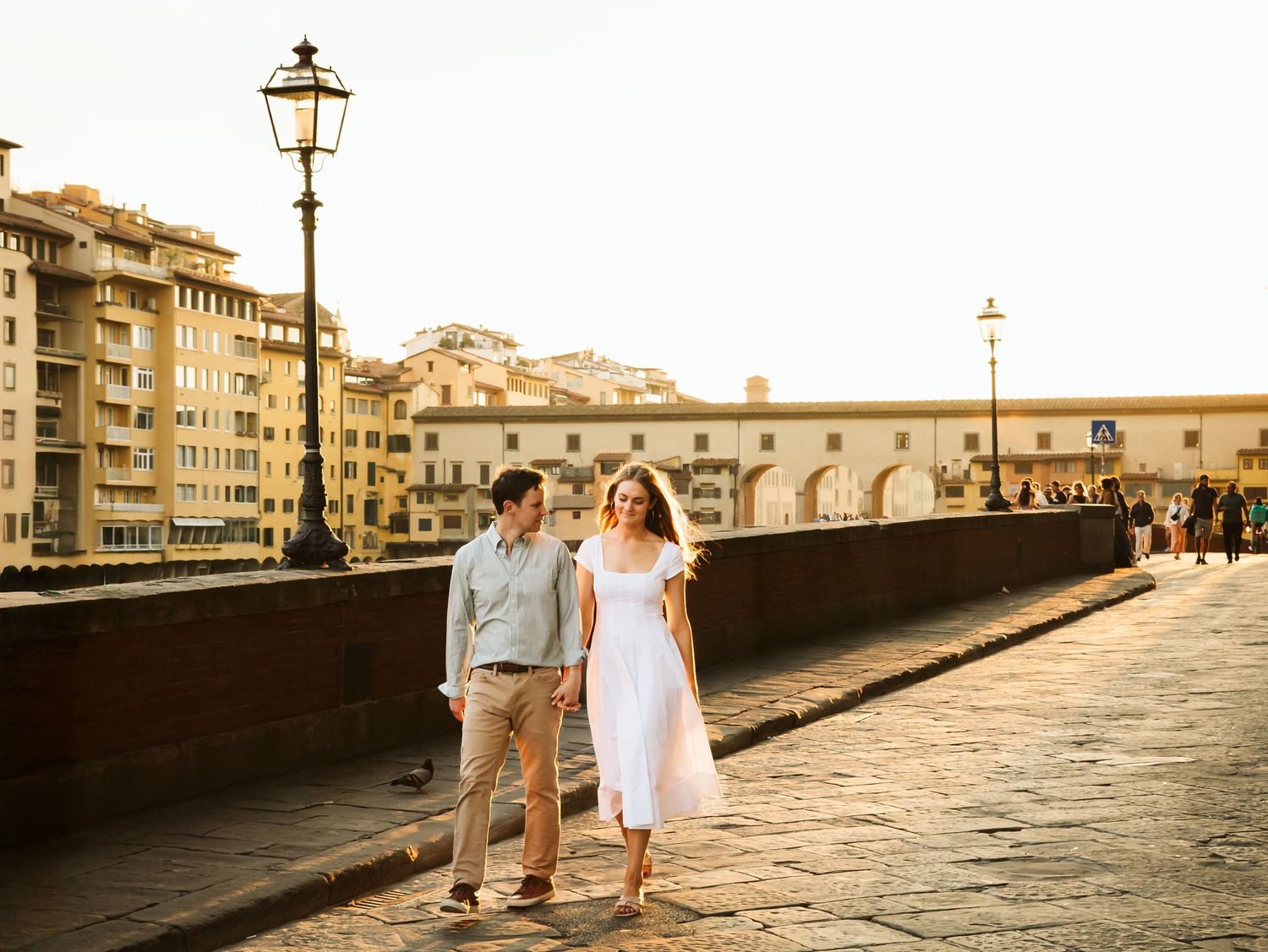 Happy couple walking hand in hand along a sunlit bridge near Ponte Vecchio in Florence.
