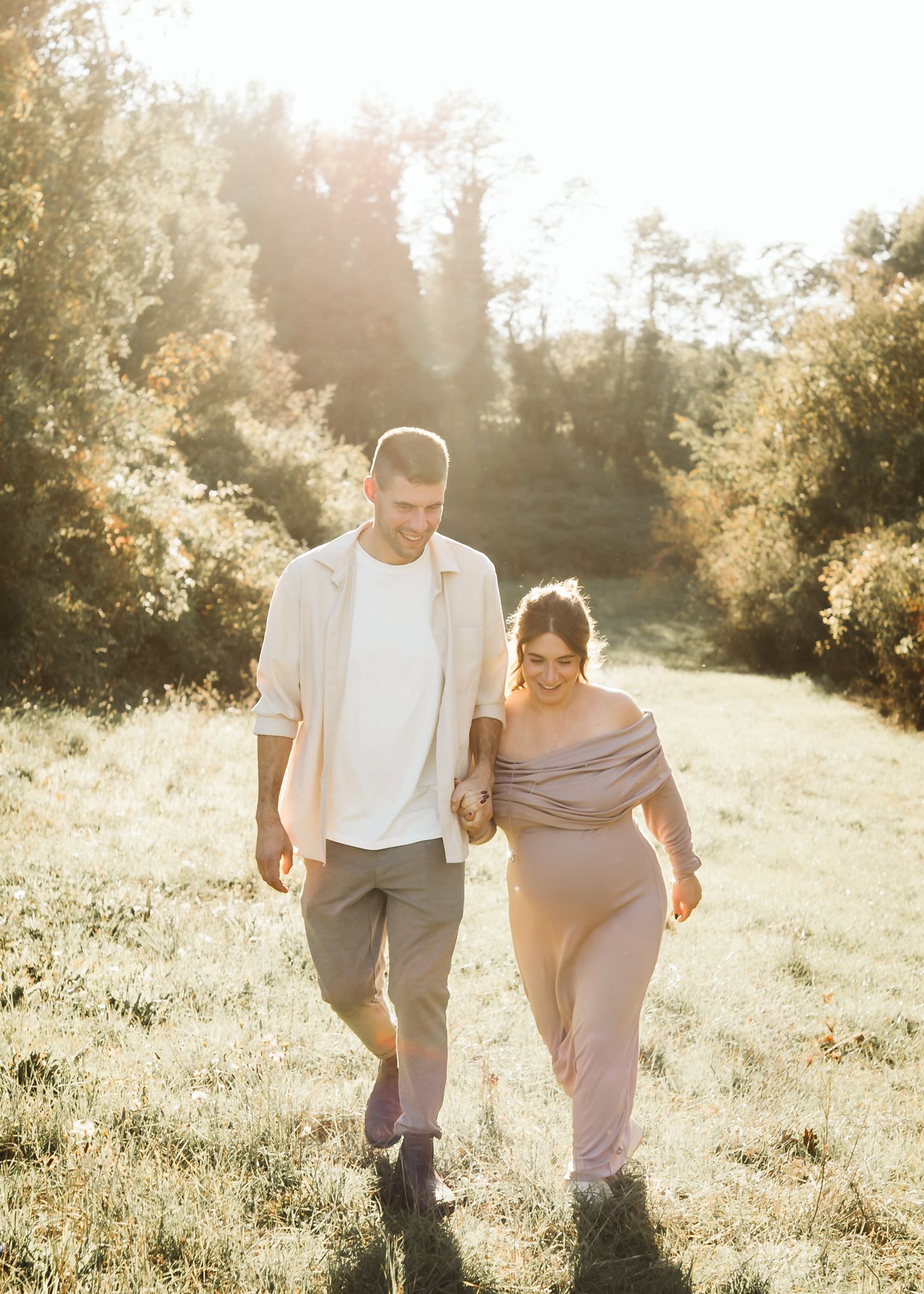 Pregnant couple walking hand in hand through a glowing sun-drenched meadow surrounded by lush greenery.