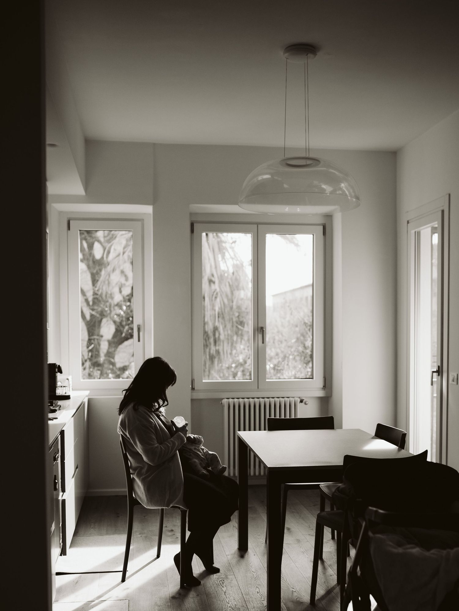 A mother sits in a sunlit kitchen nursing her newborn baby in black and white.