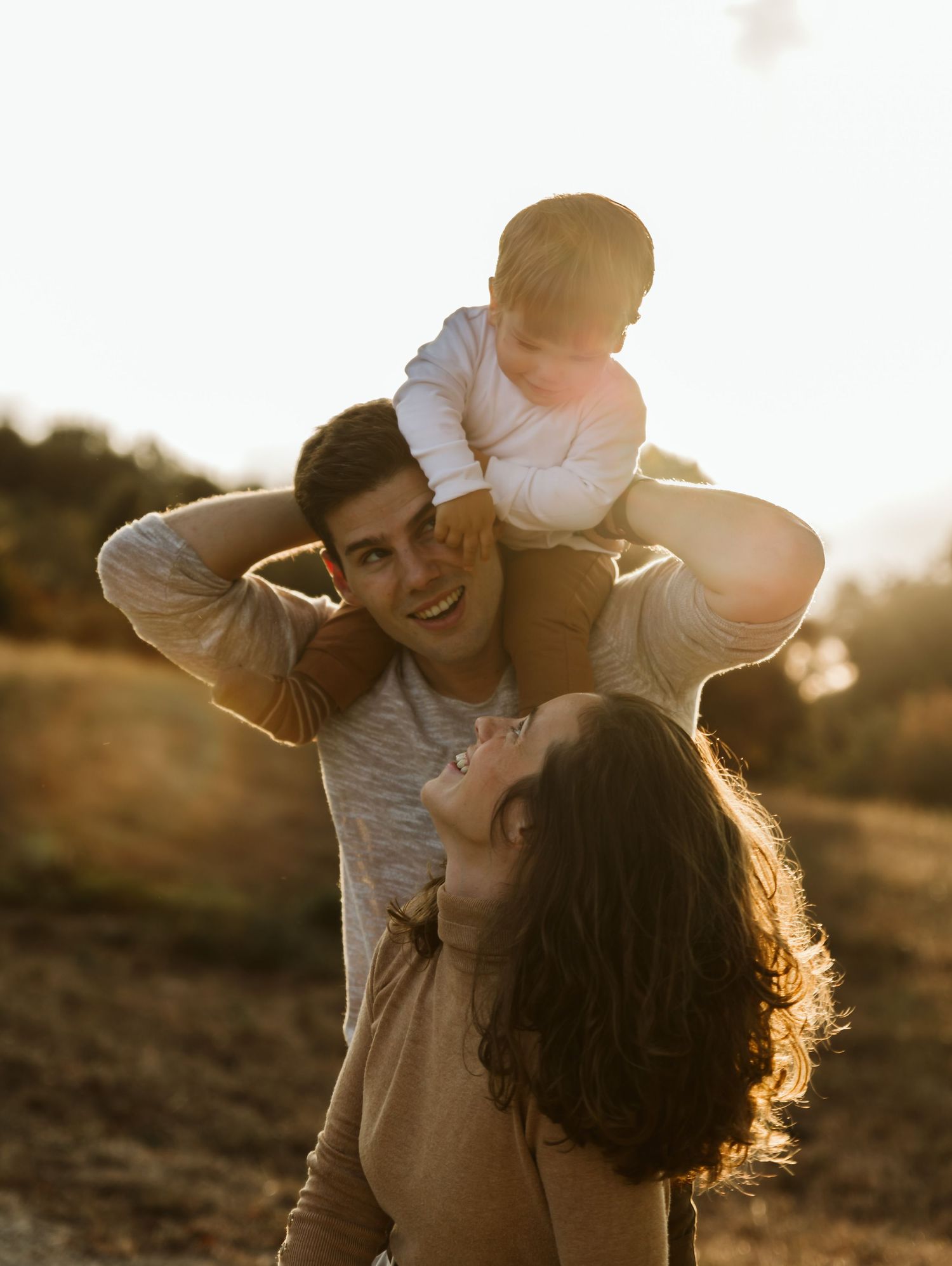 Happy parents lifting baby on shoulders in warm golden sunset light.