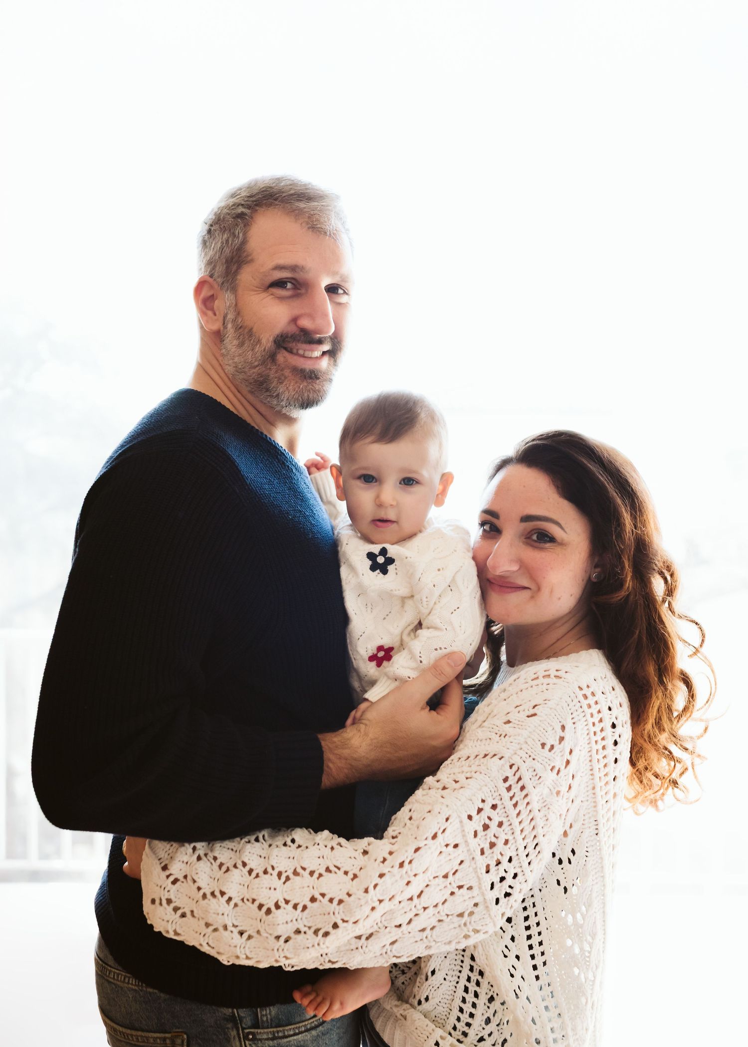 A smiling couple holds their baby girl in a bright, airy room for a family portrait.