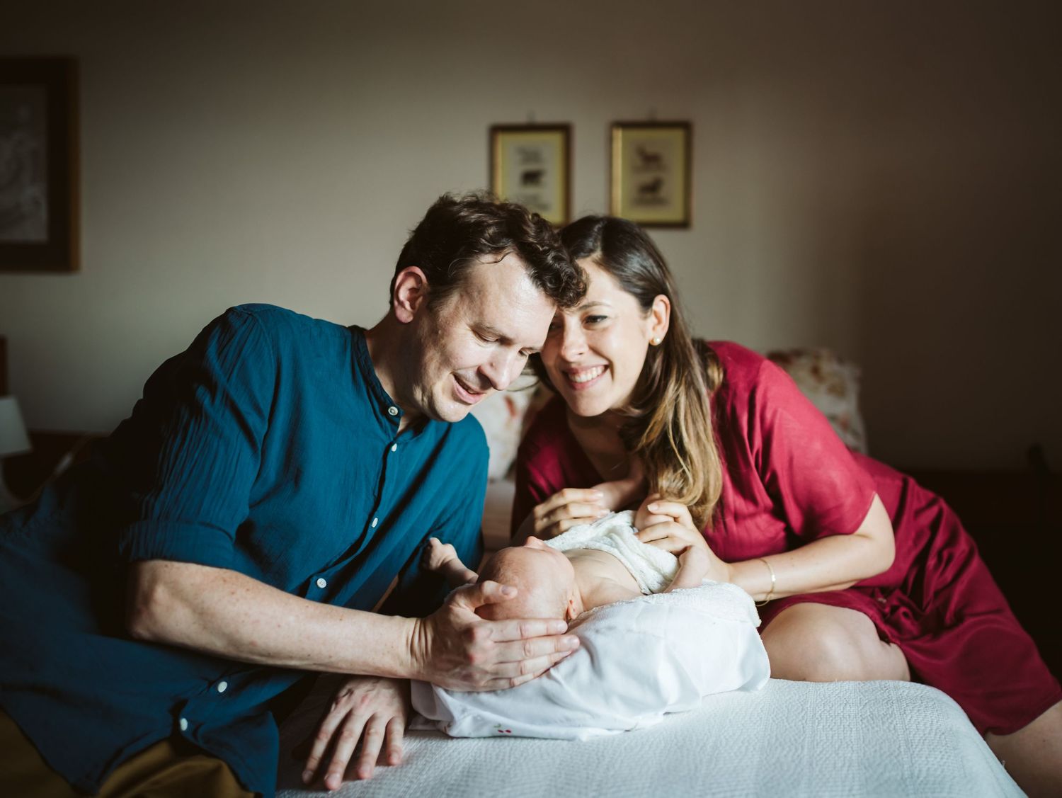 Smiling new parents sit on a bed gazing at their newborn baby wrapped in white cloth.