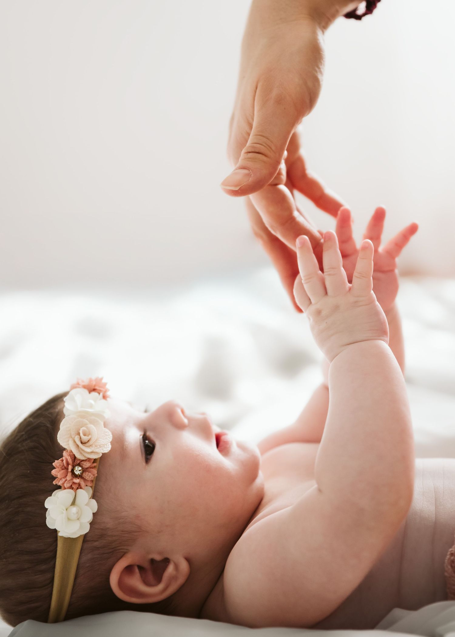 A baby girl wearing a floral headband reaches up to touch an adult's hand.