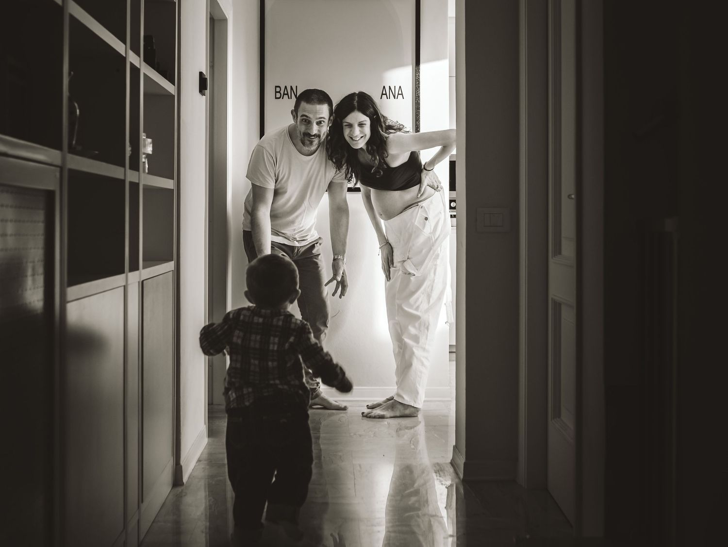 Black and white photo of parents welcoming toddler running down hallway toward them.