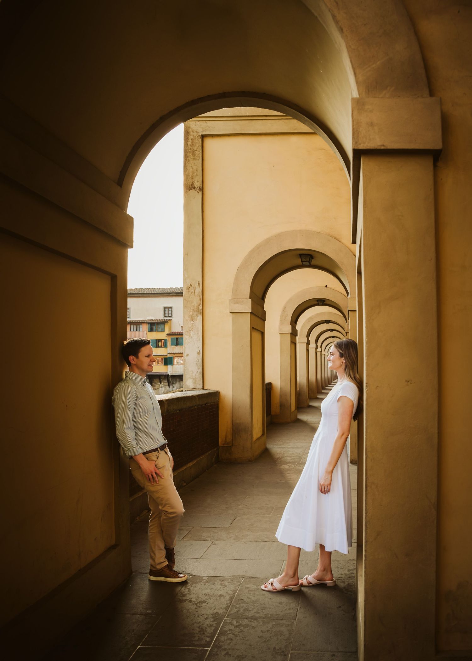 Couple standing apart in a long arched corridor overlooking Florence Italy in warm golden light.