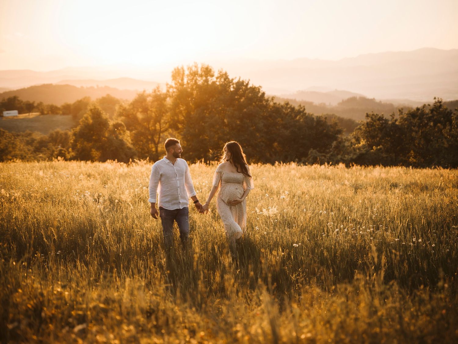 Couple holding hands walking through golden meadow at sunset with rolling hills in the background.
