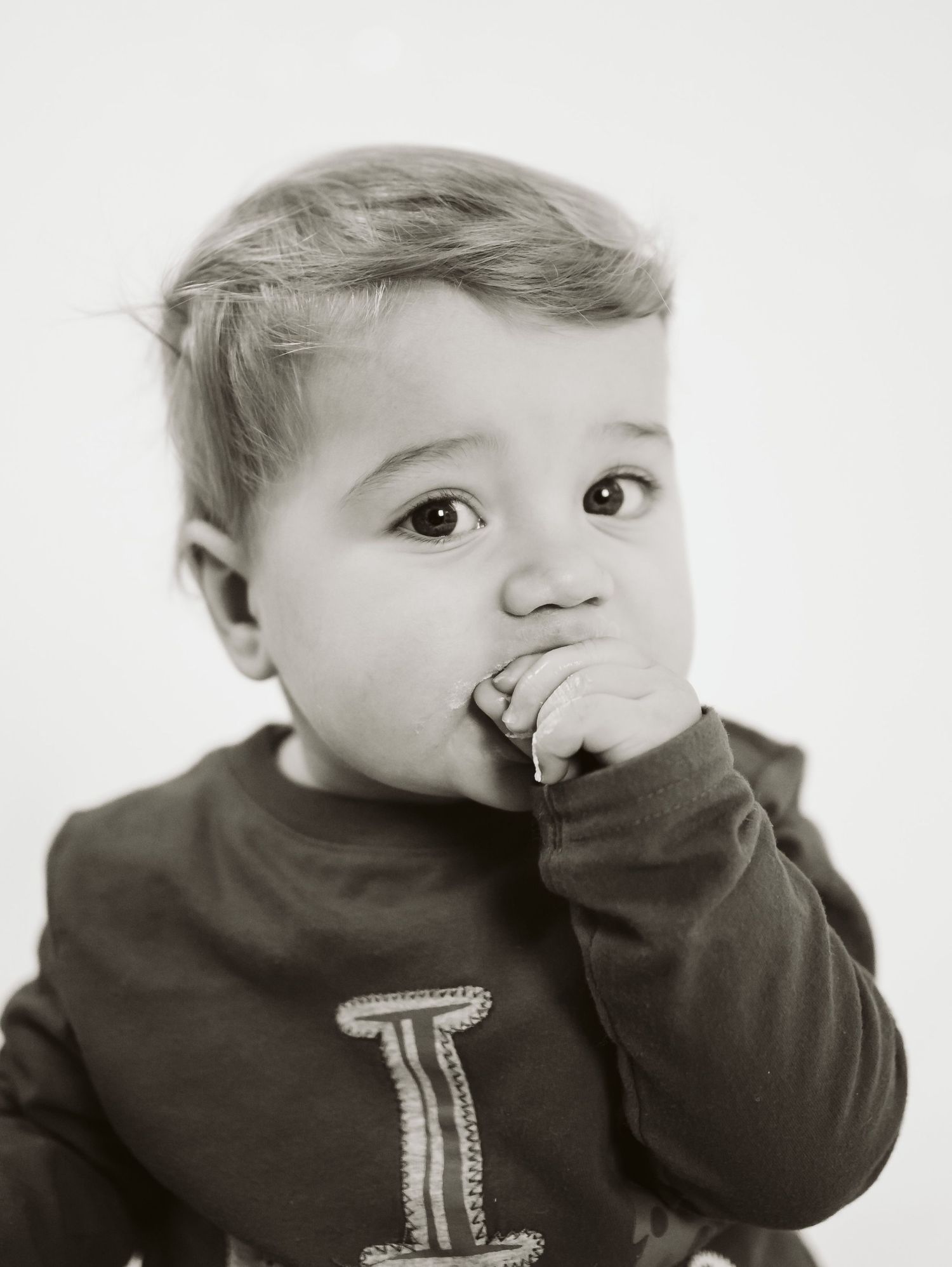 Black and white portrait of a cute toddler in a dark shirt with hand in mouth against white background.