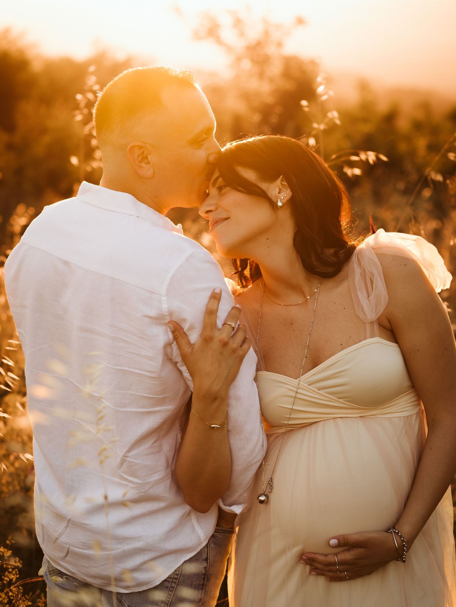 Pregnant woman in cream dress embraced by partner during golden hour outdoors.