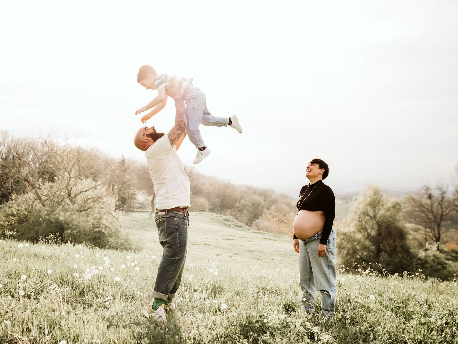 A man lifts a toddler high in the air while a pregnant woman watches in a sunny meadow.