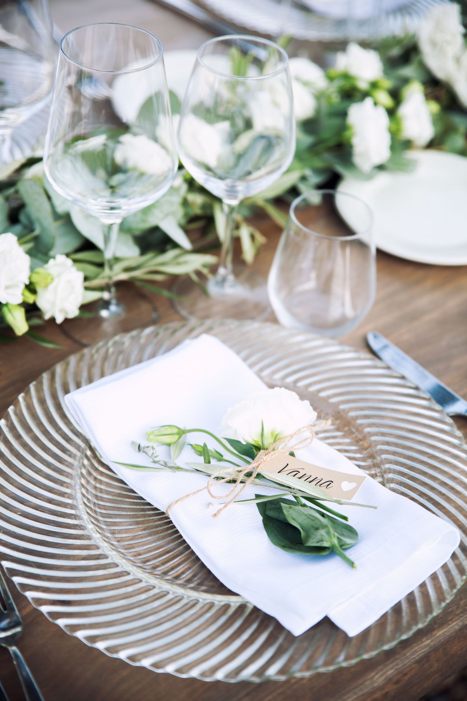 Elegant wedding table setting with glass charger plate, white napkin, name card, and floral decor.