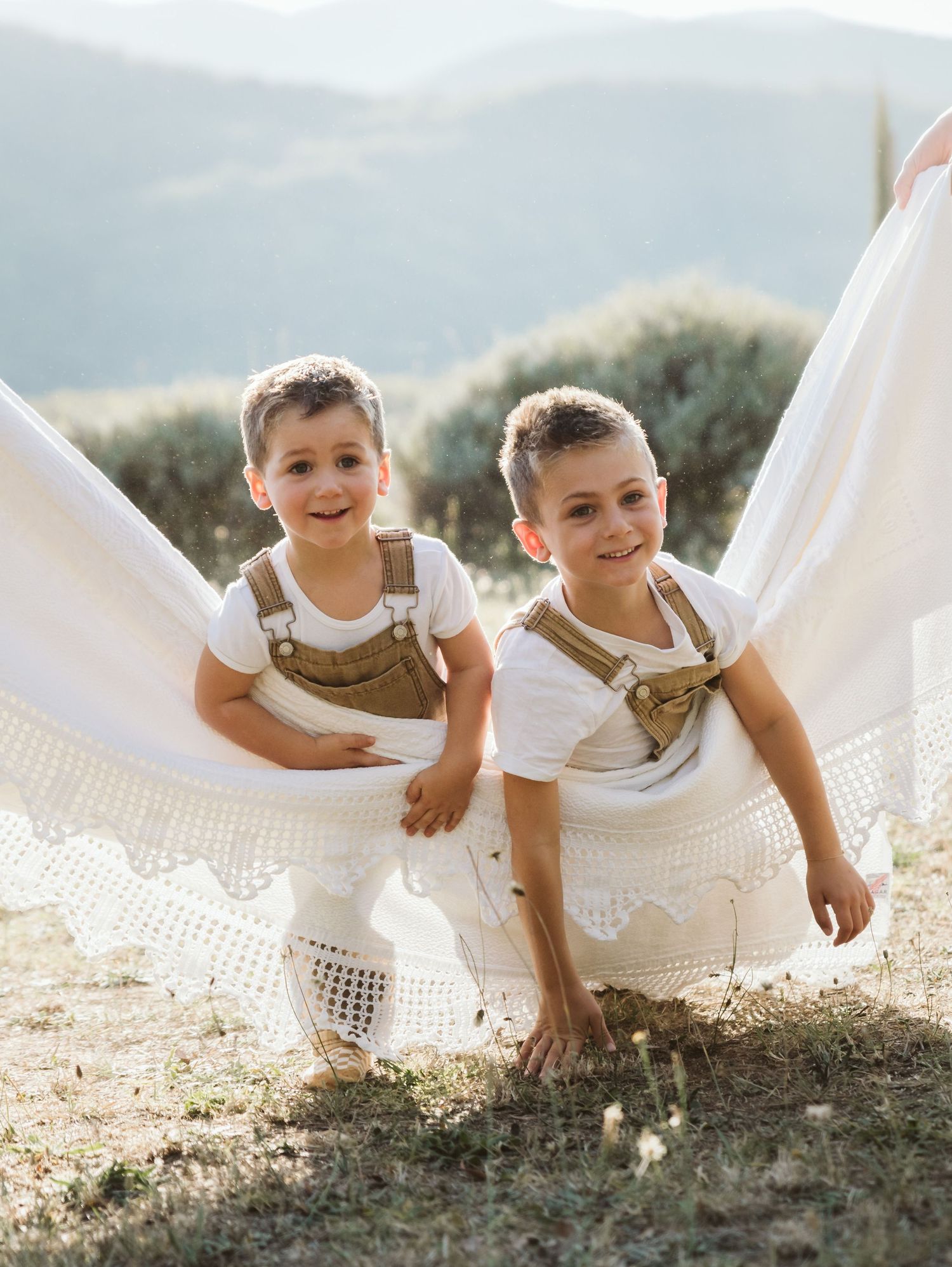 Twin boys in matching brown overalls and white shirts posing with flowing white fabric outdoors.