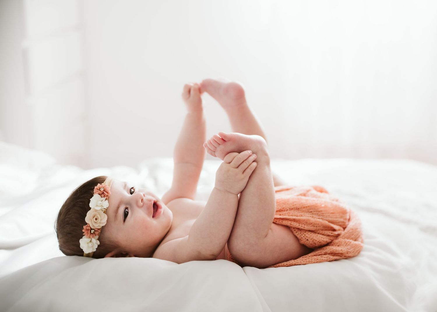 Adorable baby girl in orange bloomers and floral headband lying on white bed.