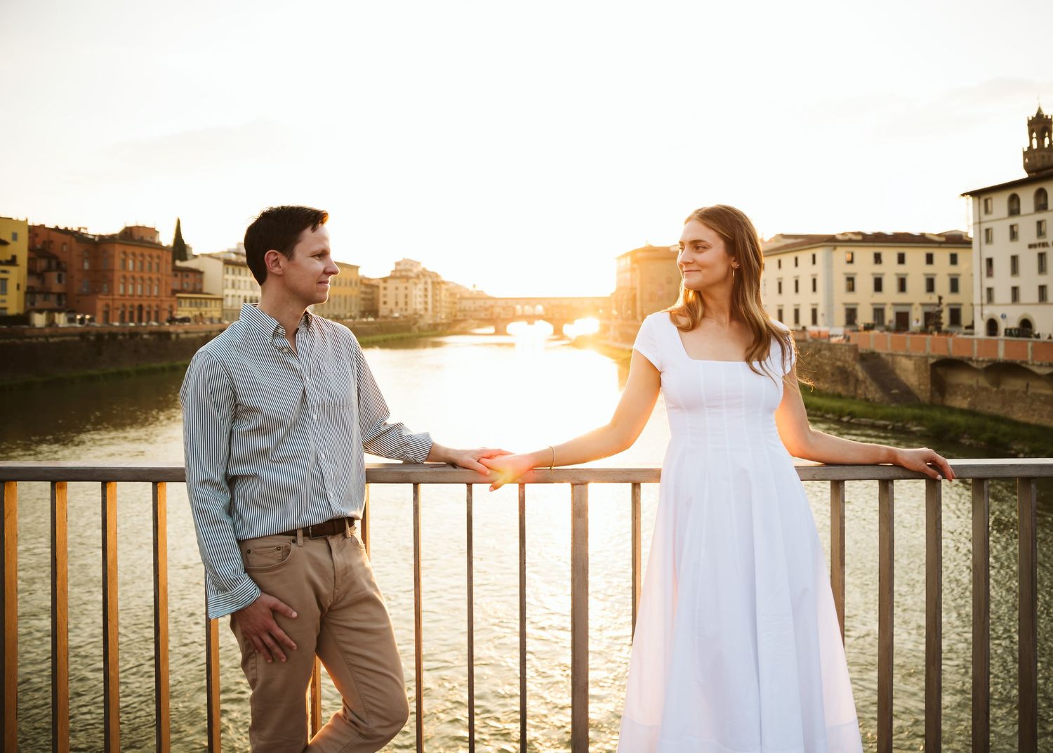 Couple holding hands on bridge with golden sunset and Arno River in Florence.