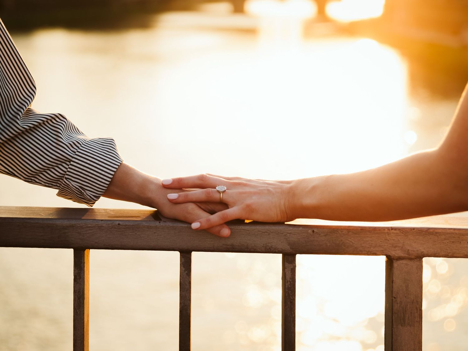 Couple holding hands on a railing by a sunlit river at golden hour.