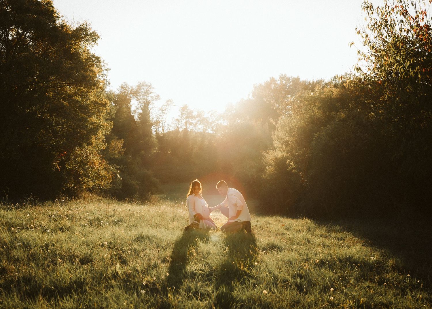 Romantic couple sitting together in a sunlit meadow surrounded by lush green trees at golden hour.