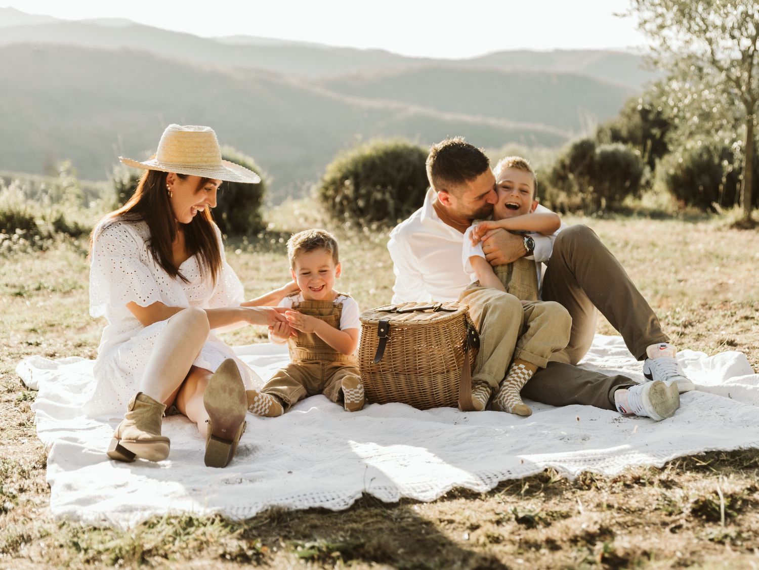 Family enjoying a picnic on a white blanket in a sunny mountain meadow.