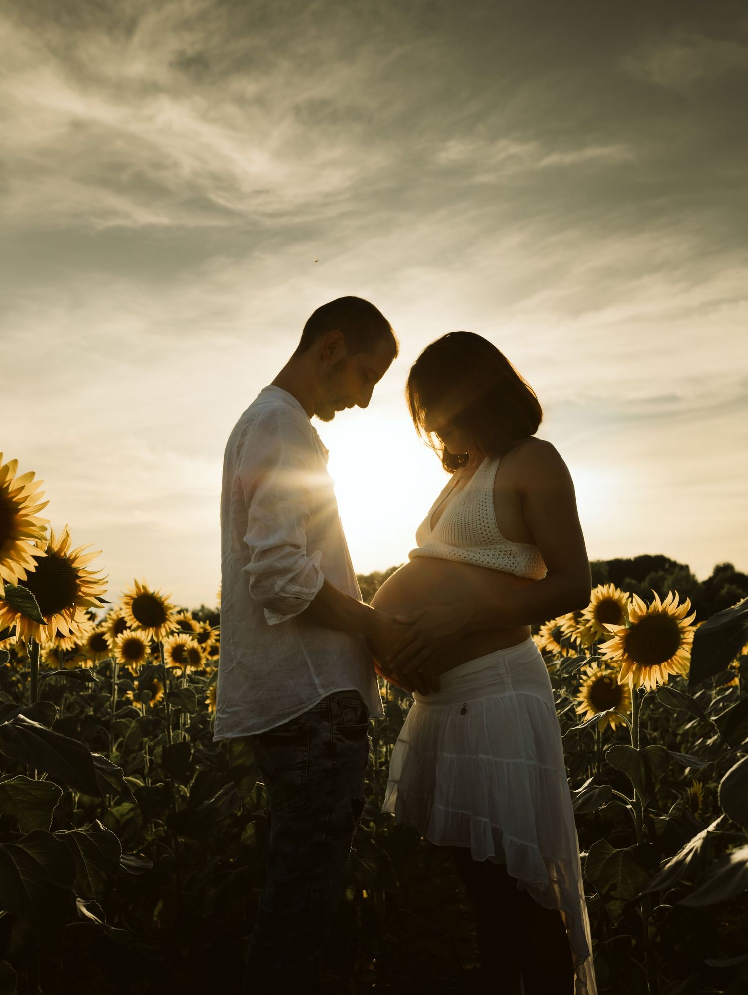 Expectant couple holding baby bump in sunflower field at golden sunset.