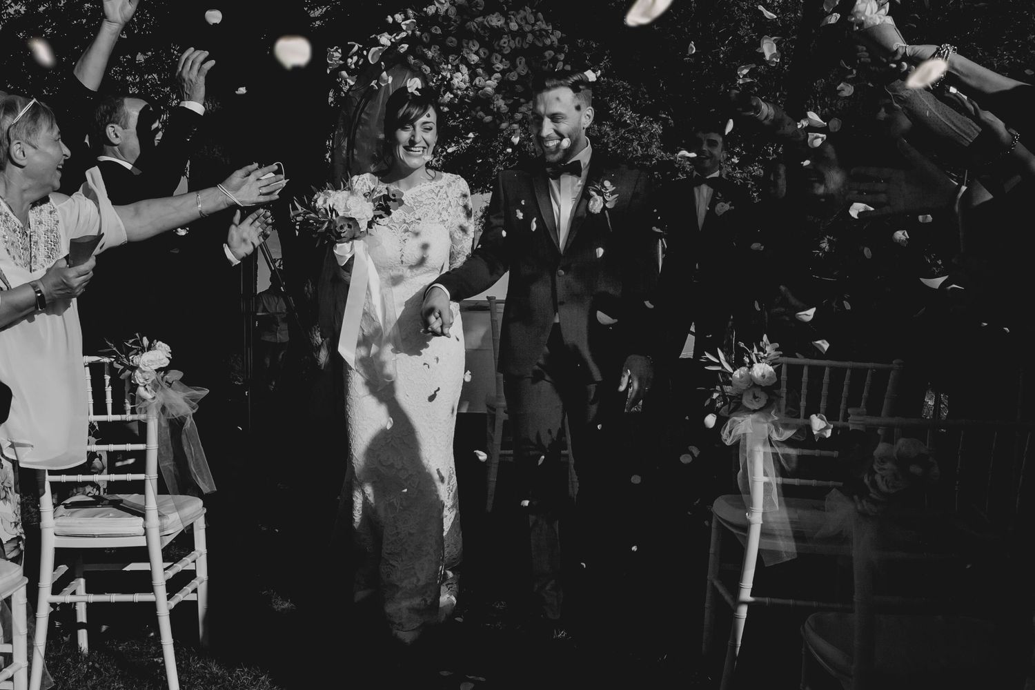 Black and white photo of newlyweds walking through a joyful confetti shower at their outdoor ceremony.