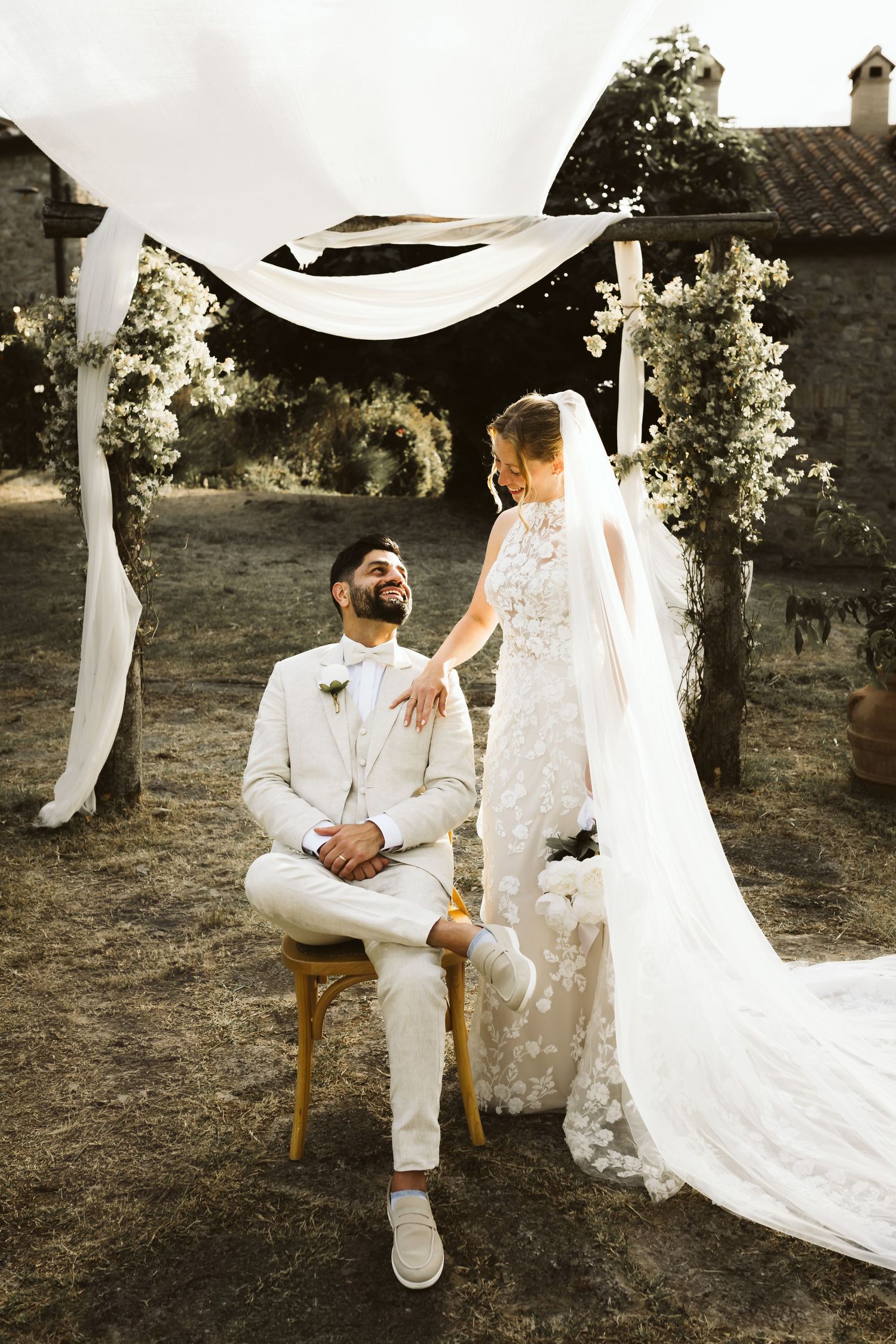 Groom seated smiling at bride in lace wedding dress under white draped arch.