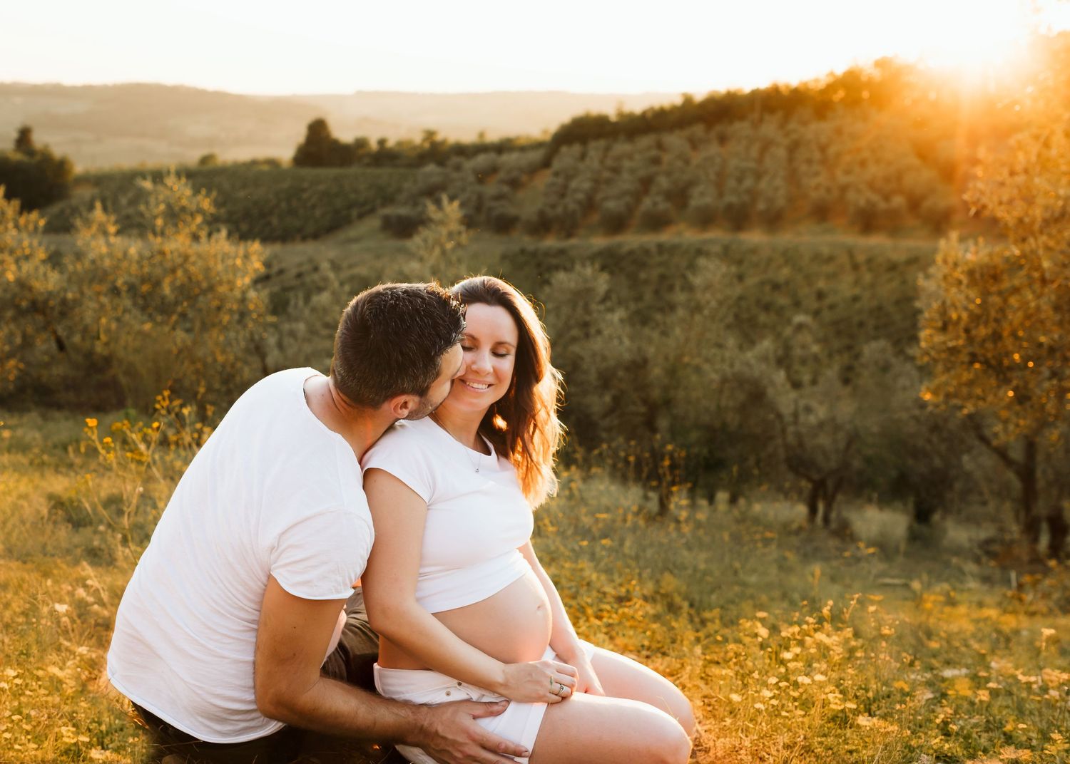 Happy pregnant couple sitting together in a sunny field at golden hour, man kissing womans cheek.