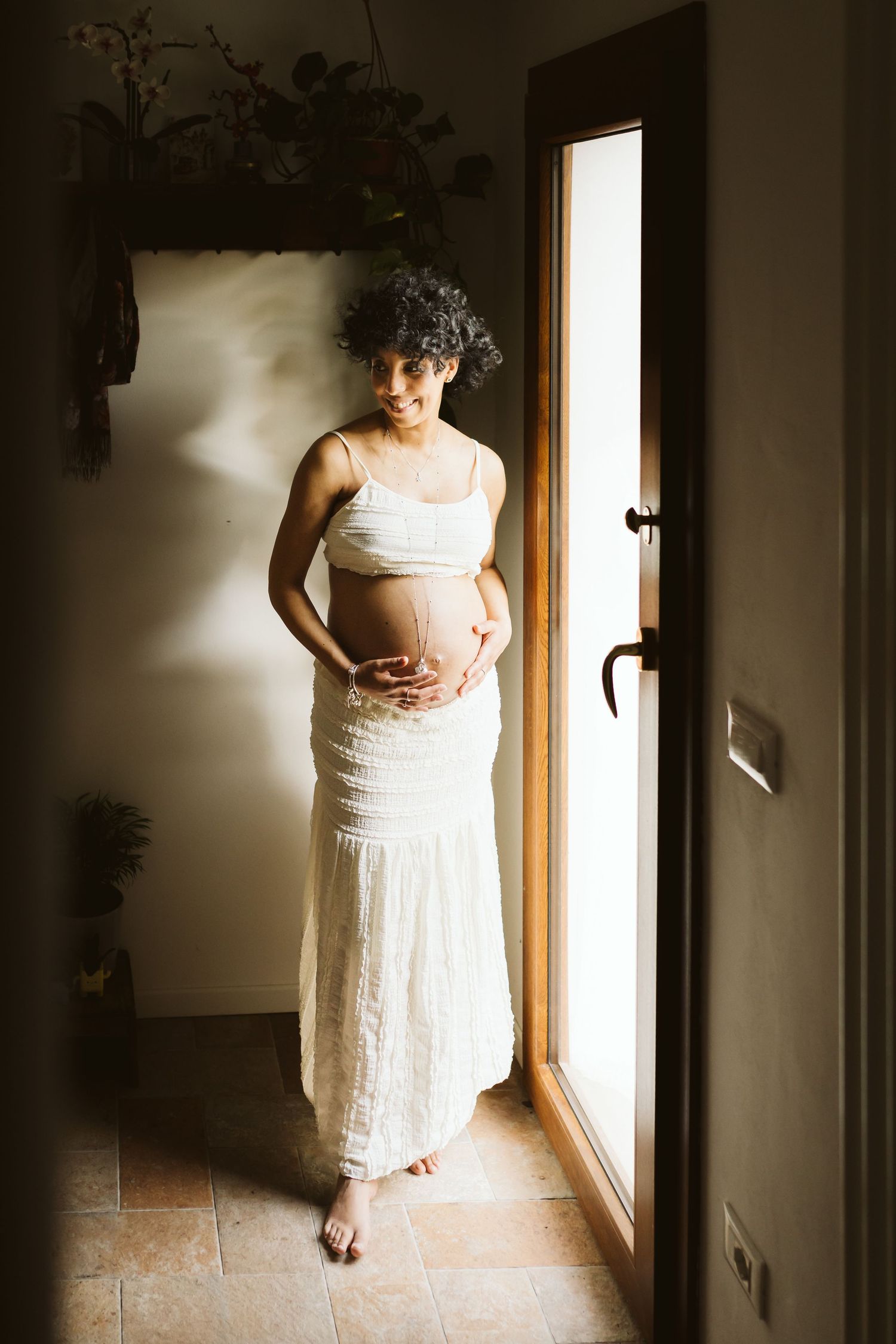 Smiling pregnant woman in white dress standing by doorway in natural light.