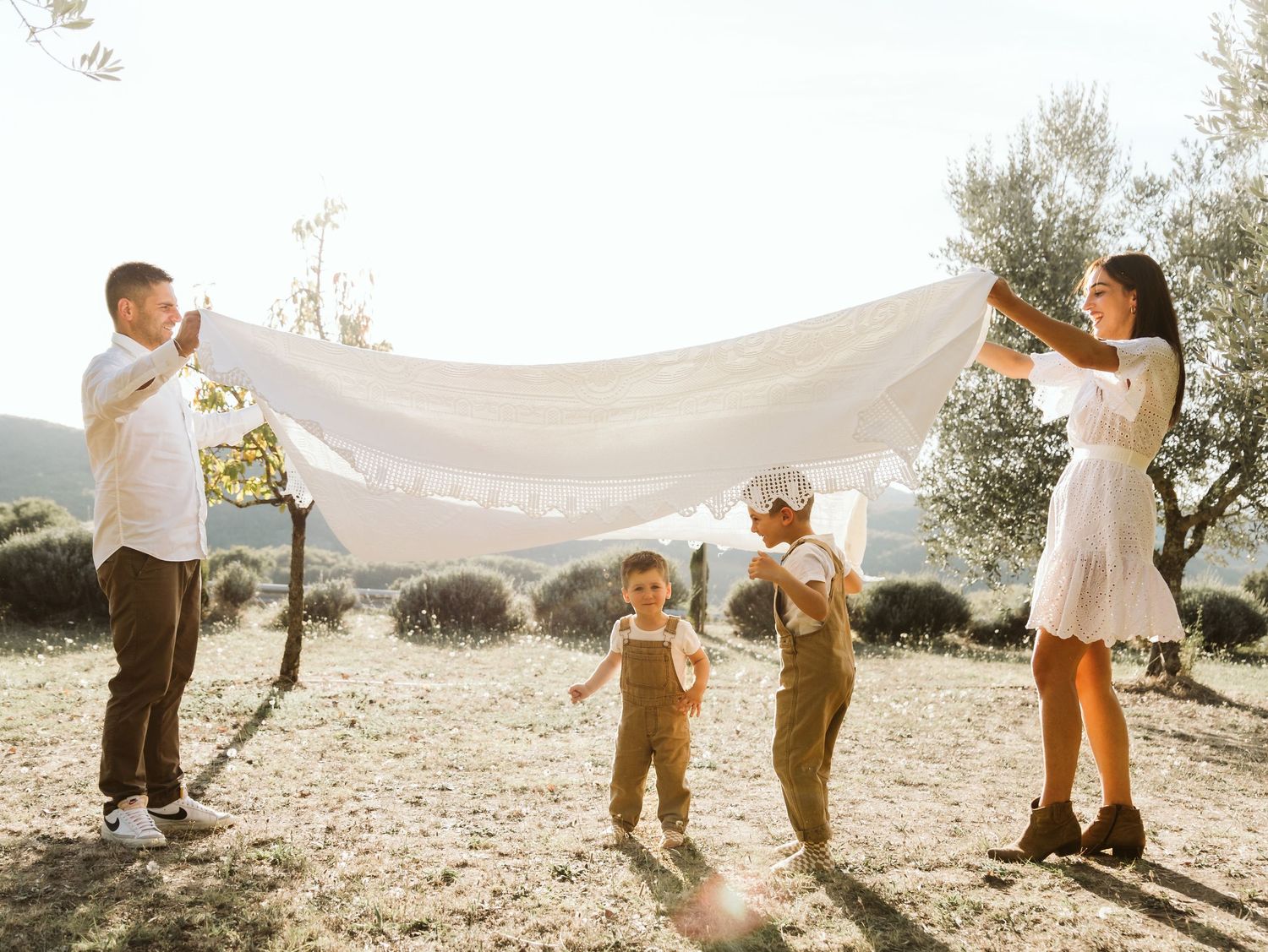 Family of four playfully holding a white sheet outdoors in a sunny olive grove.