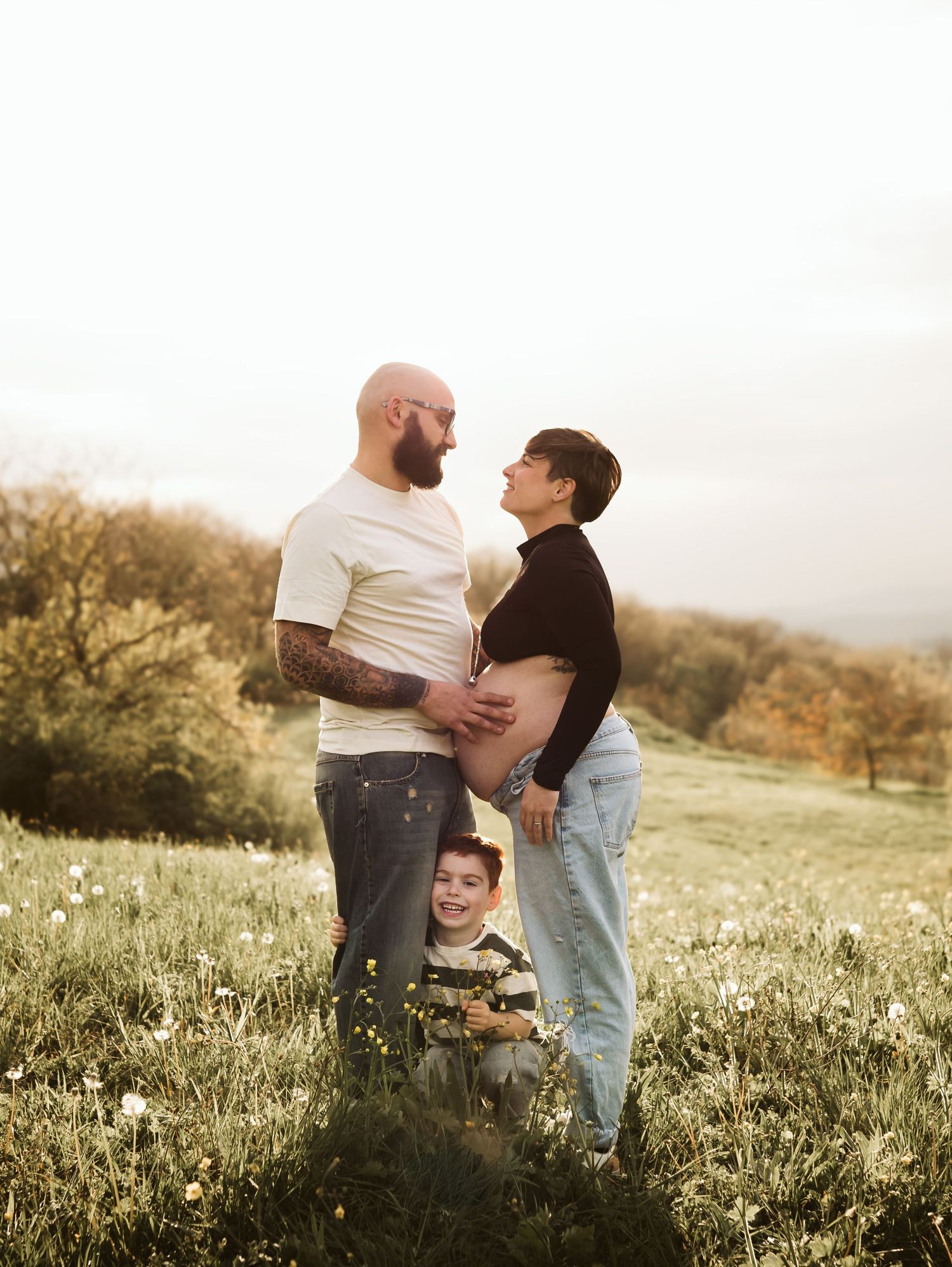 Tattooed father, pregnant mother, and young son posing together in a blooming meadow.
