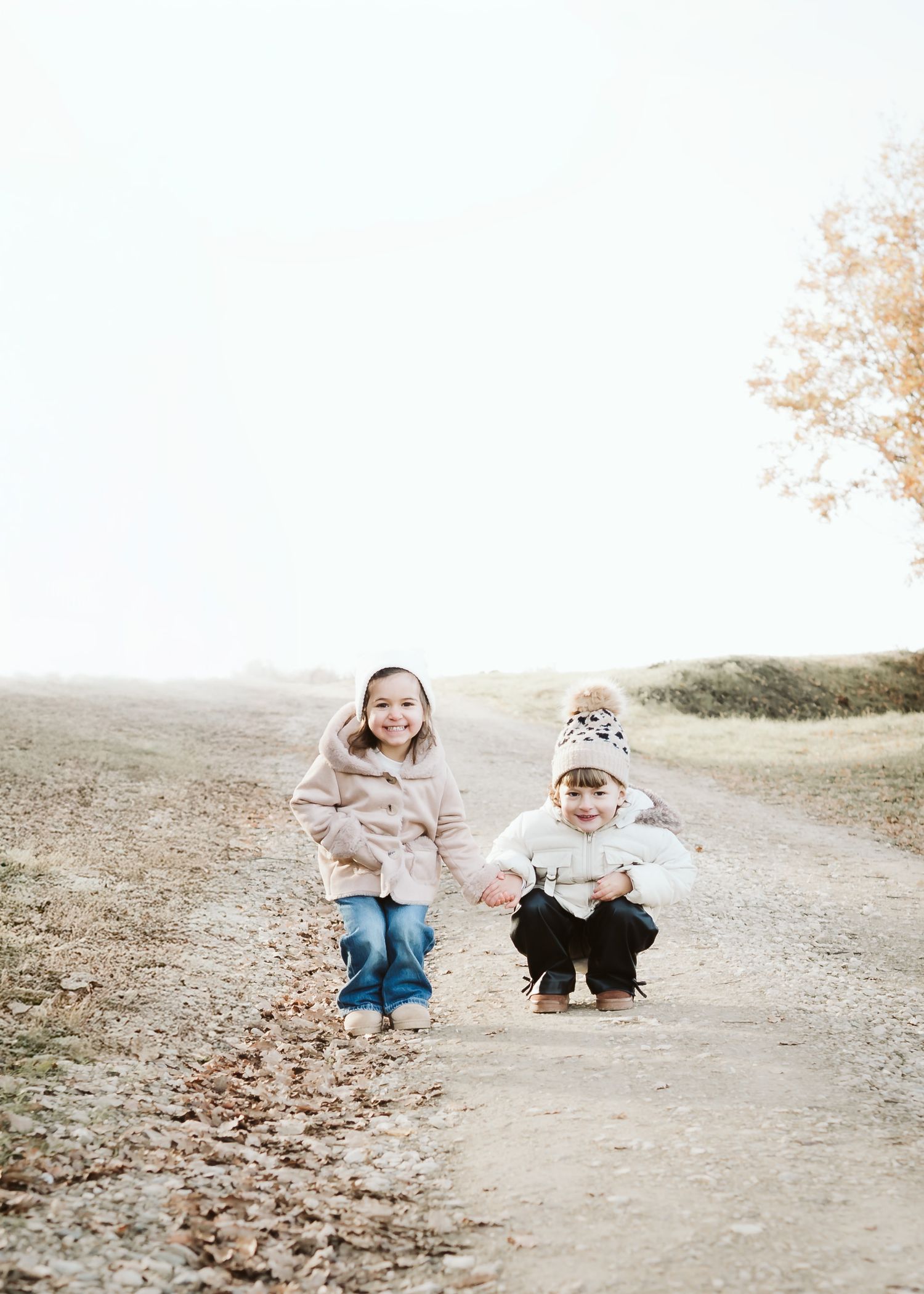 Two young siblings hold hands on a dirt path surrounded by autumn leaves on a misty day.