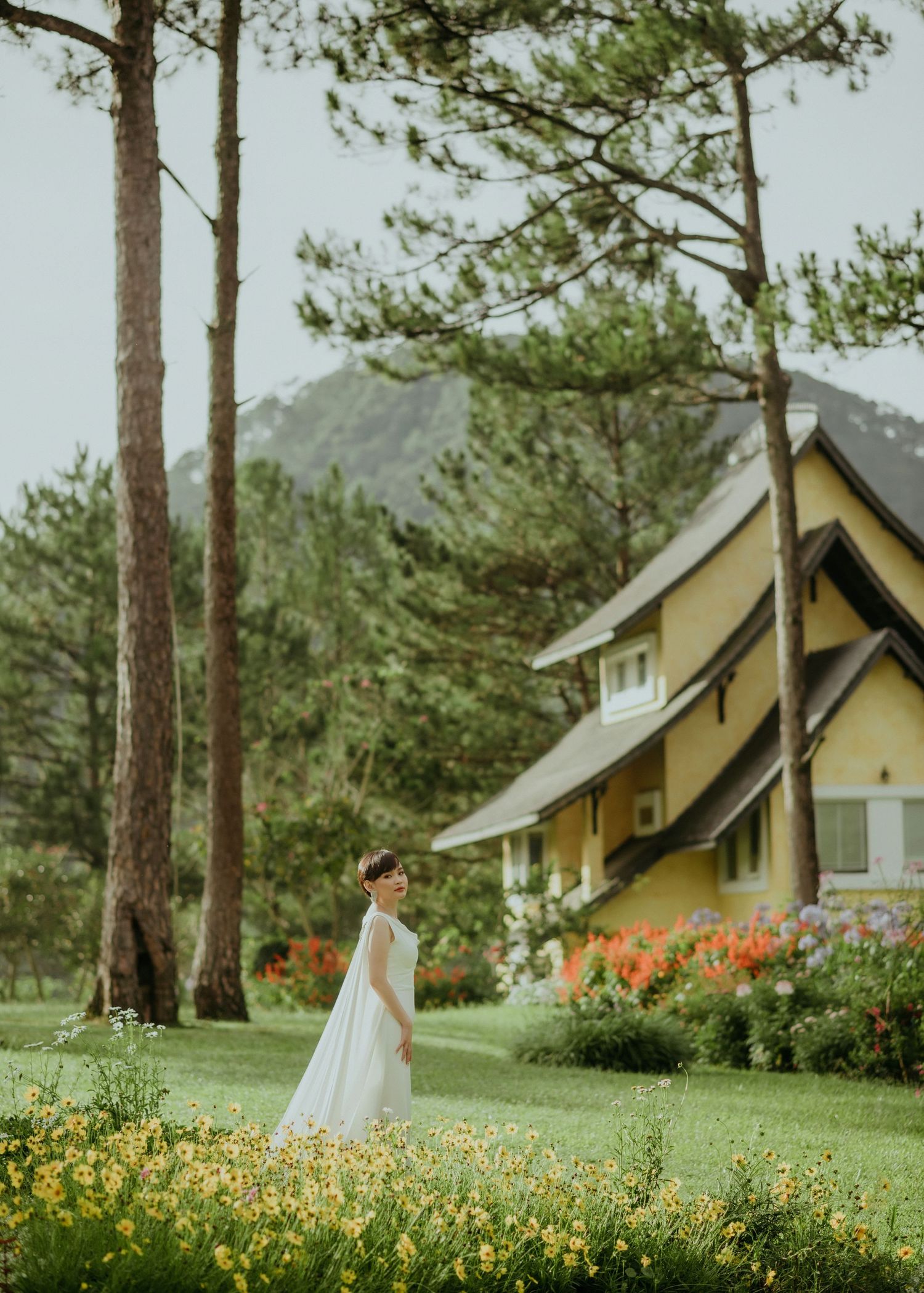 A yellow cottage style house nestled among tall pine trees with vibrant flowers blooming in the foreground.