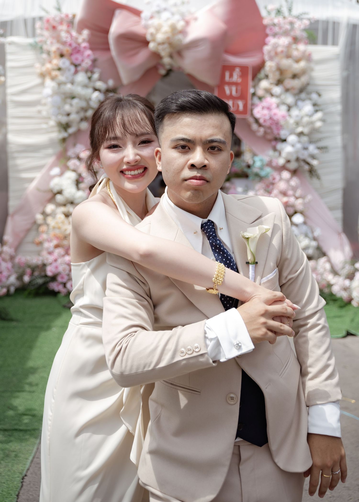 A couple poses together in front of a pink and white floral arch with a large bow decoration.