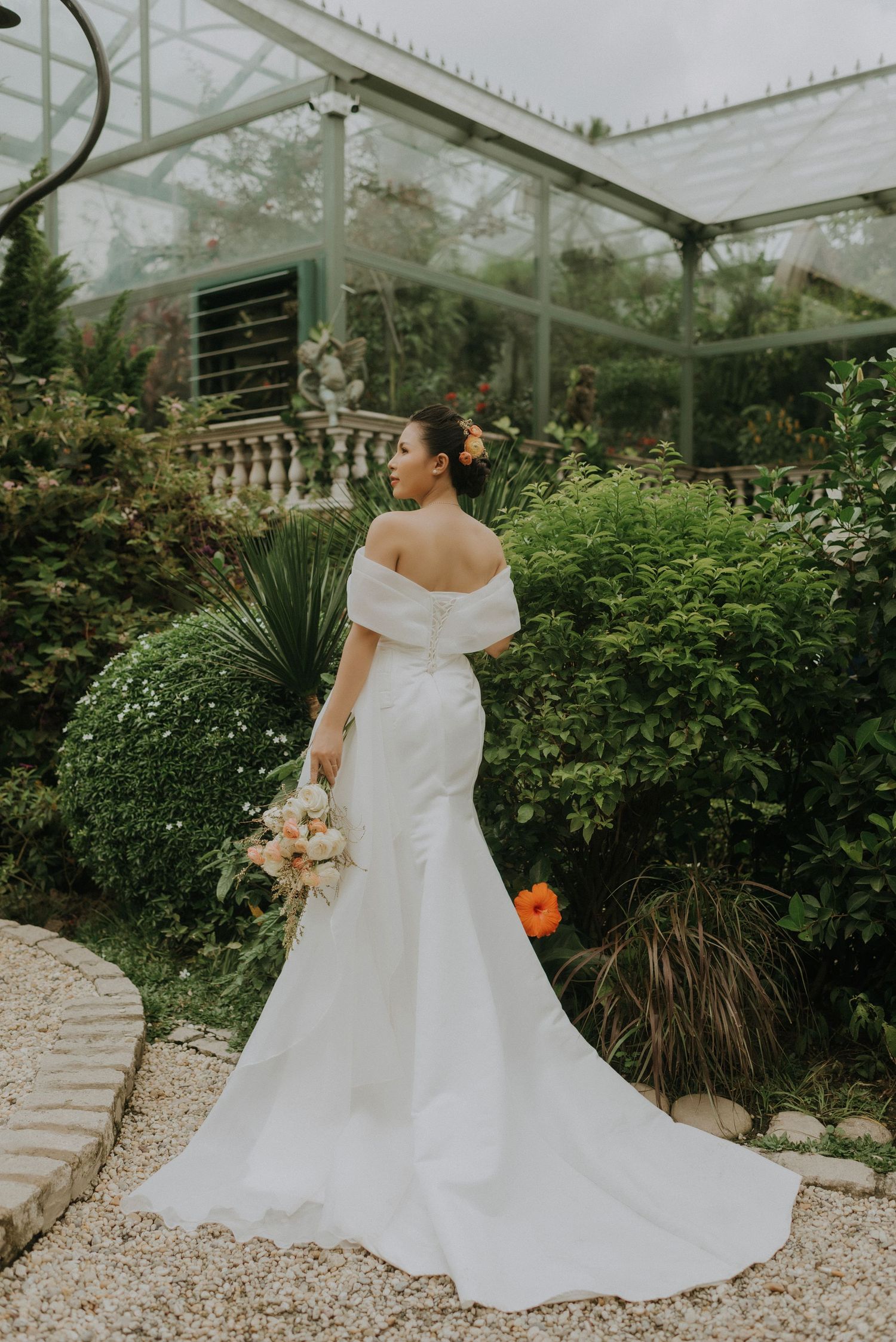 Off-shoulder white wedding gown with long train photographed in a lush greenhouse garden setting.