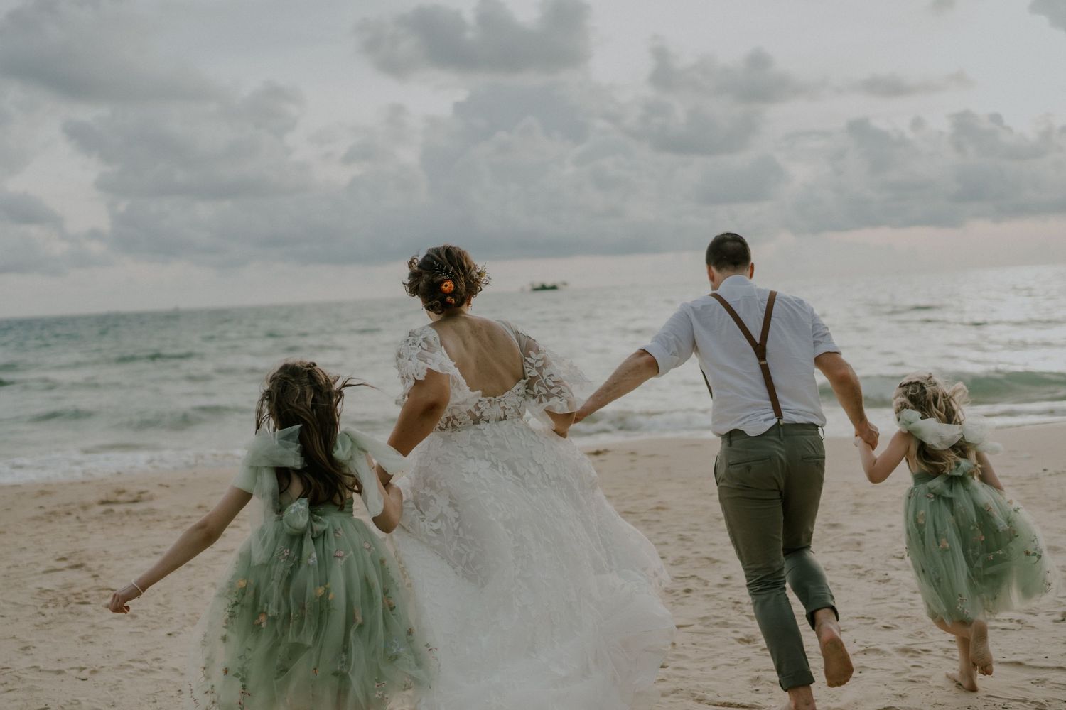 Family running joyfully on beach with bride in white dress and children in sage green outfits.