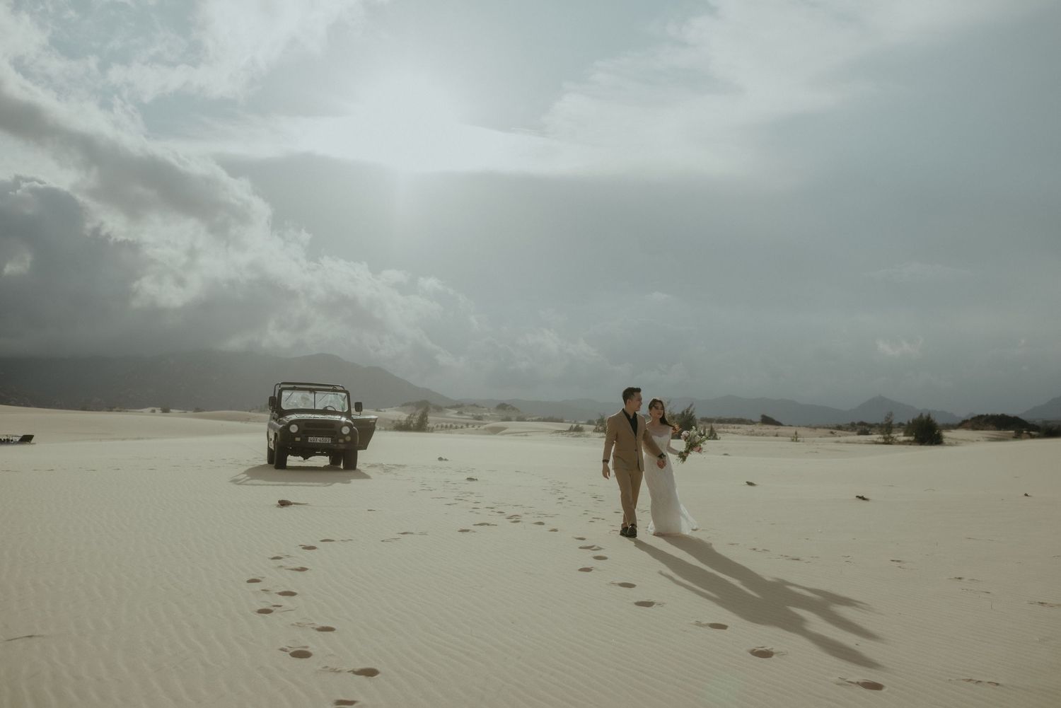 Two people walk across a pristine white sand desert with their footprints trailing behind and a vehicle in the distance.