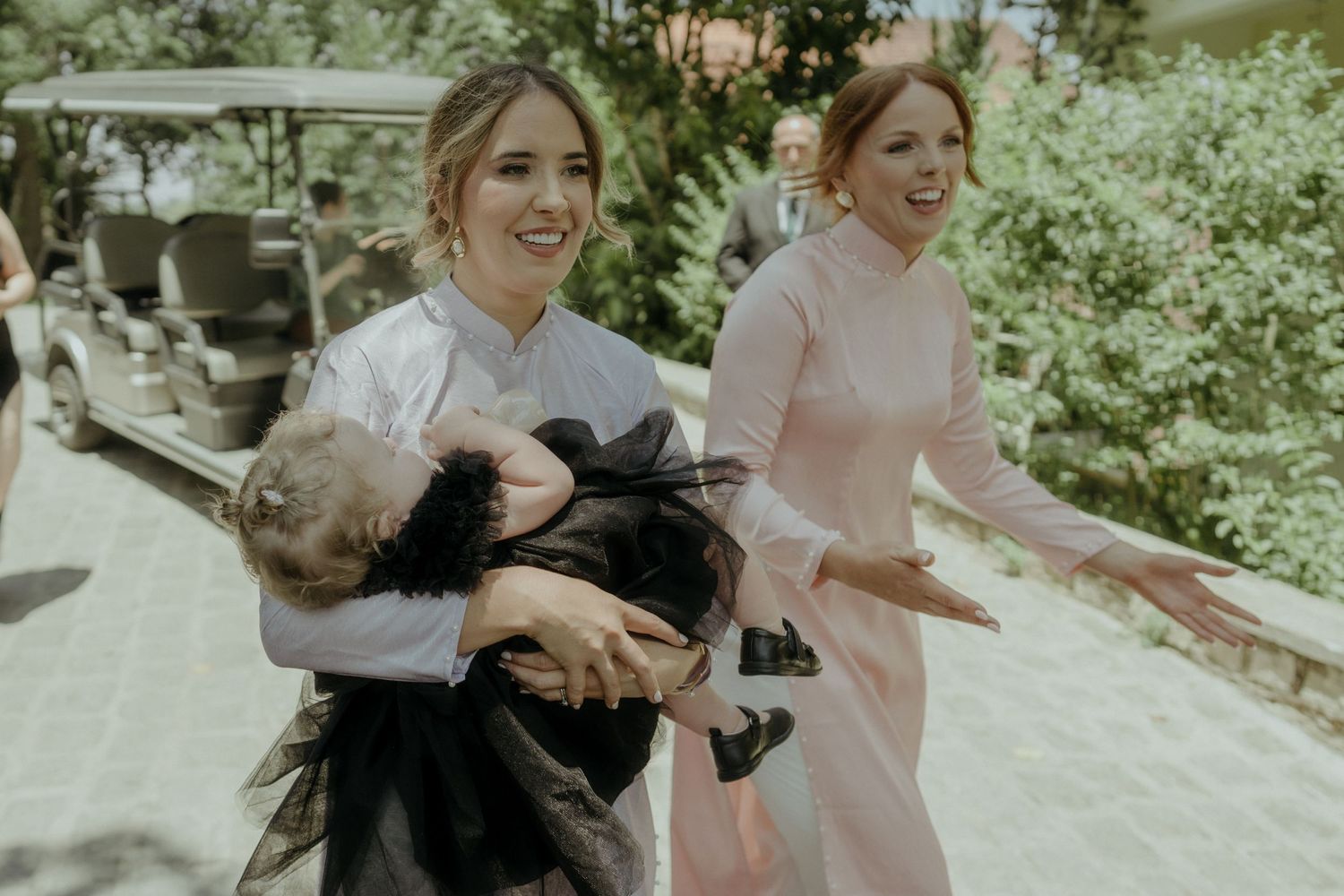 Two stylishly dressed women in pastel outfits share a joyful moment while holding a fluffy black dog outdoors.