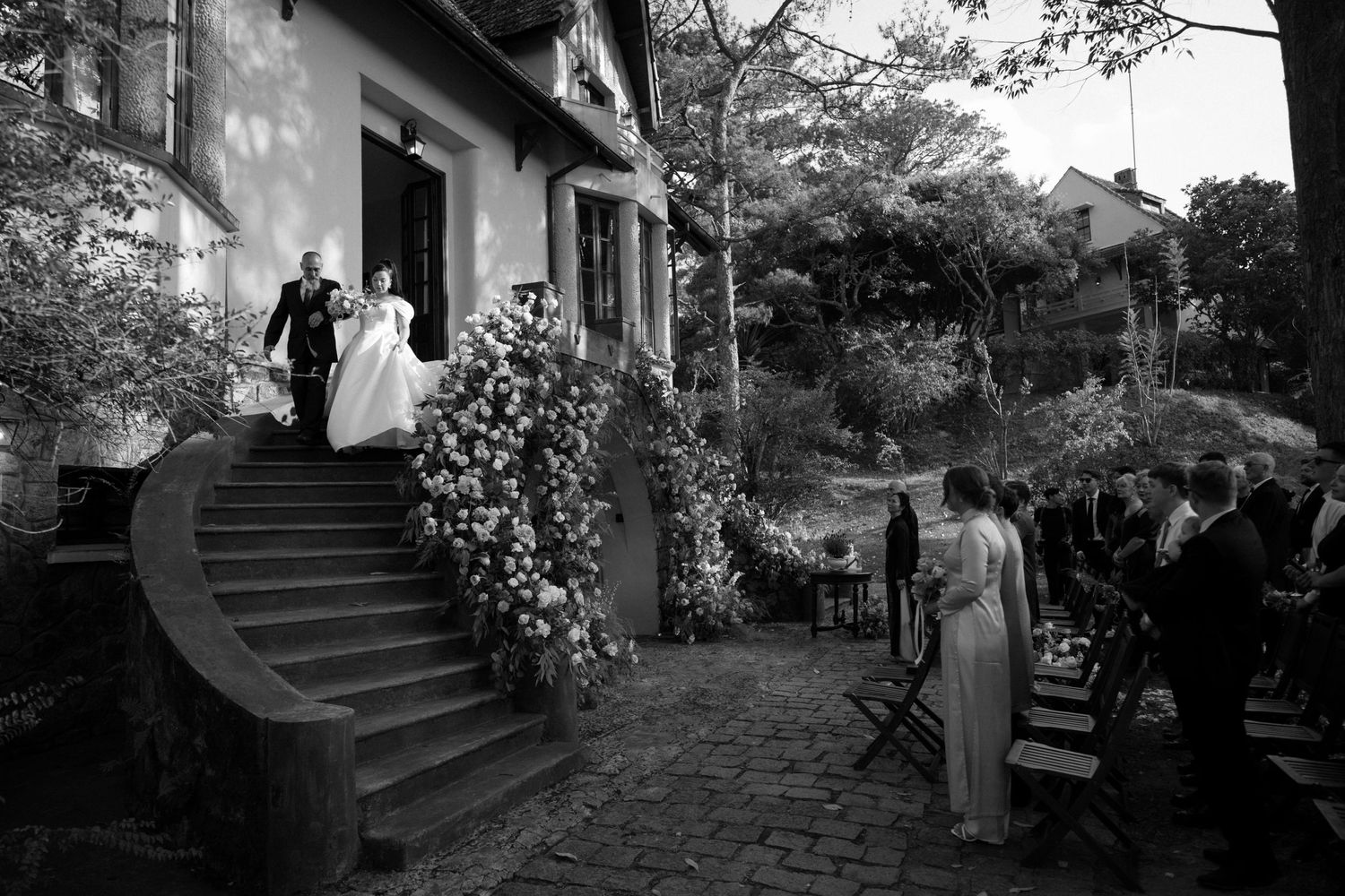A black and white wedding photograph showing elegantly dressed people gathered on curved garden steps of a house.