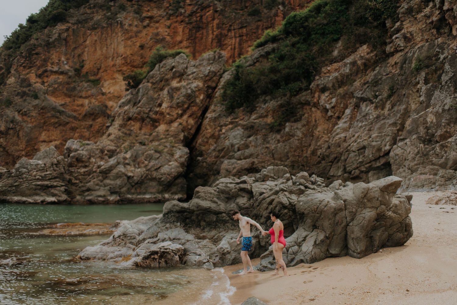 Two people exploring rocky beach formations near orange cliffs.