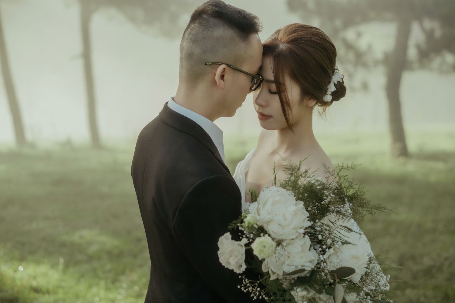 Romantic wedding photo of a couple embracing in a misty forest setting with white floral bouquet.