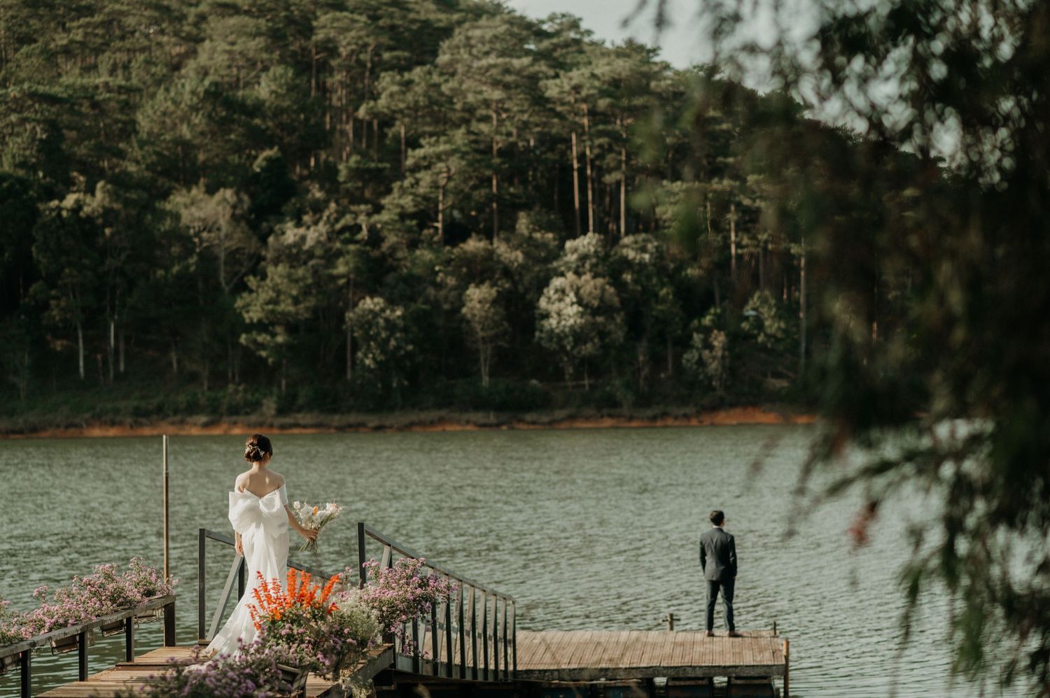 Two people stand at opposite ends of a wooden dock on a serene lake surrounded by lush forest and mountains.