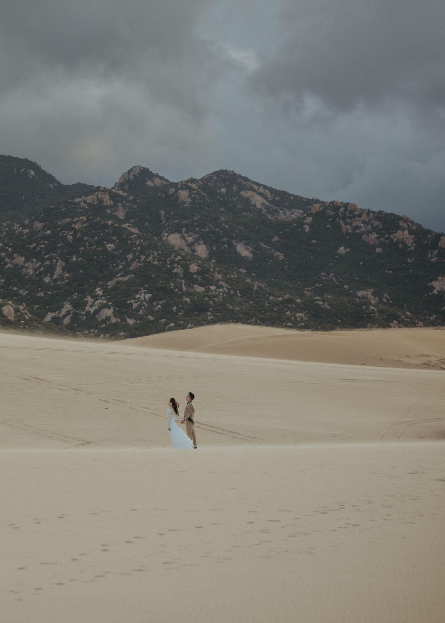 Two figures walking across vast sand dunes beneath dramatic mountain peaks and stormy skies at Great Sand Dunes National Park.