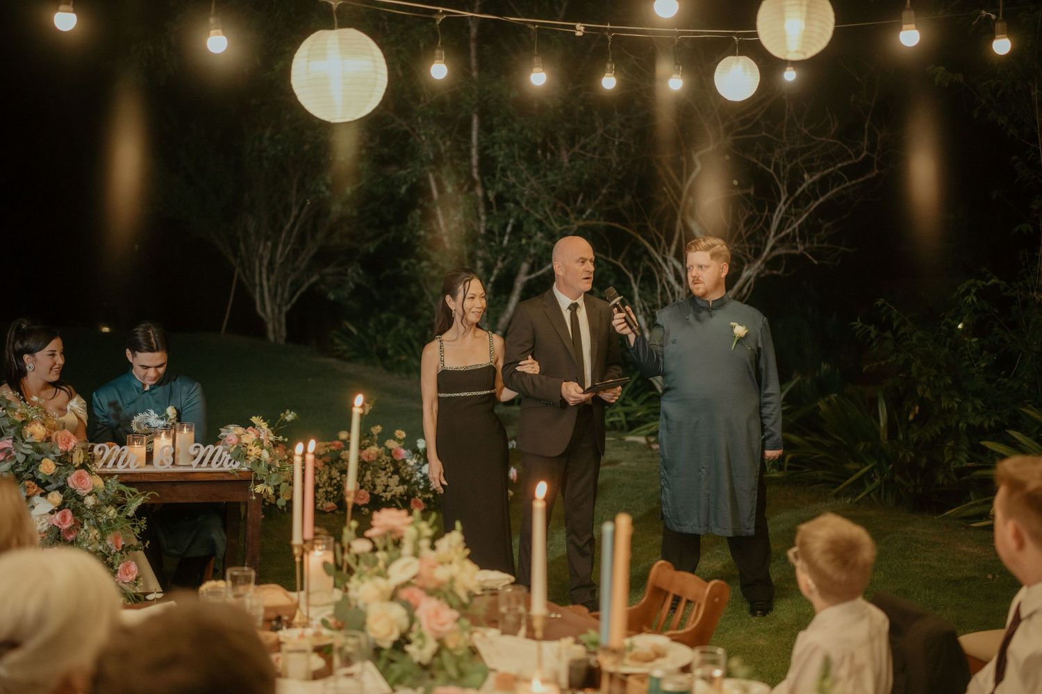 An outdoor evening wedding reception with string lights and paper lanterns illuminating a decorated dinner table setting.