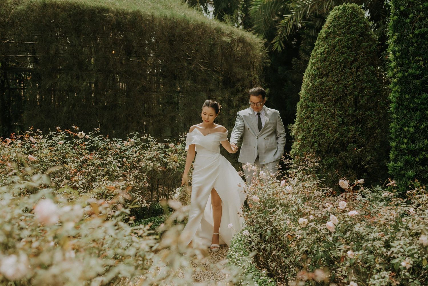 A couple walks hand in hand through a lush garden with blooming flowers and manicured hedges on their wedding day.
