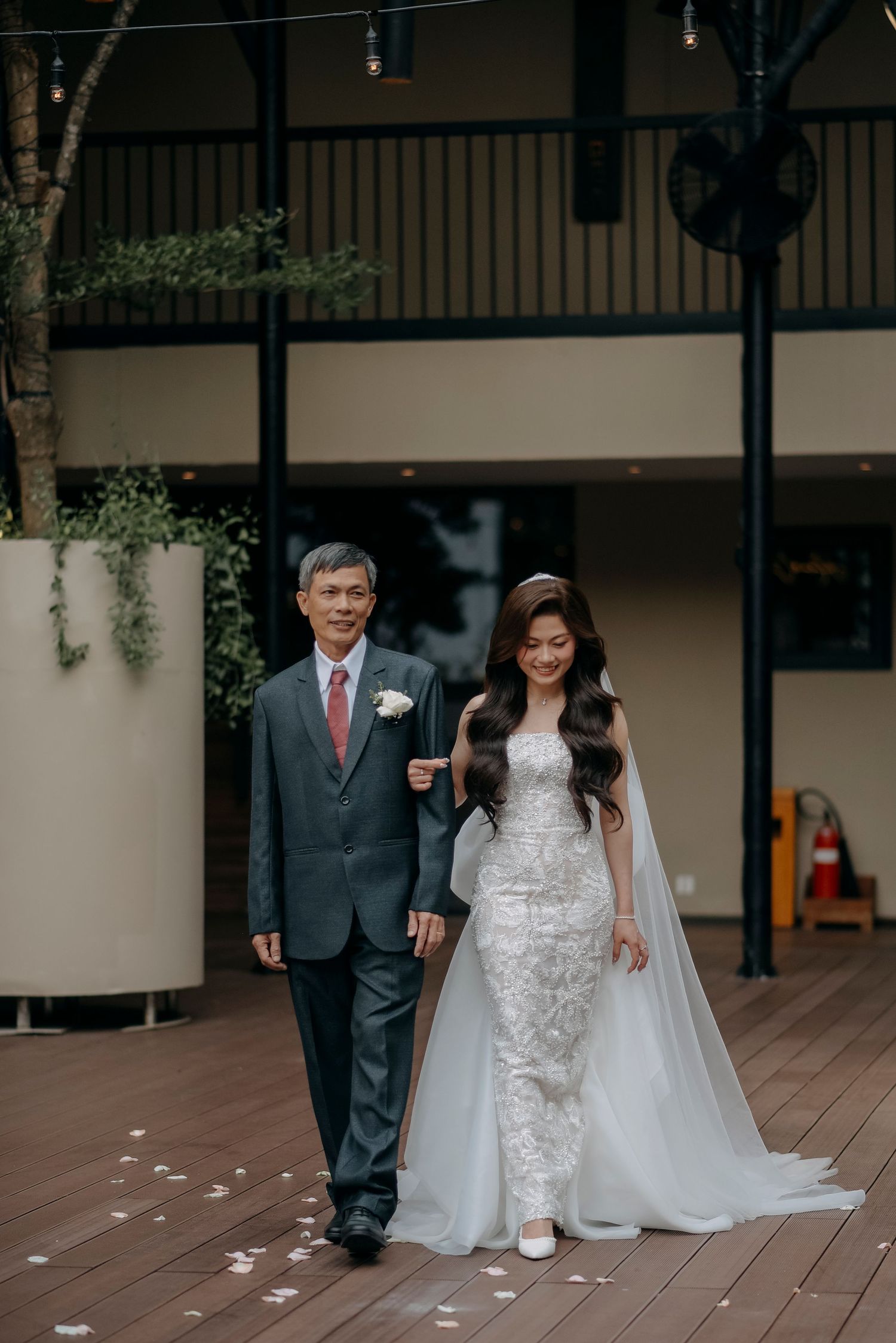 A bride in lace gown and long veil walks with her escort on a wooden deck.