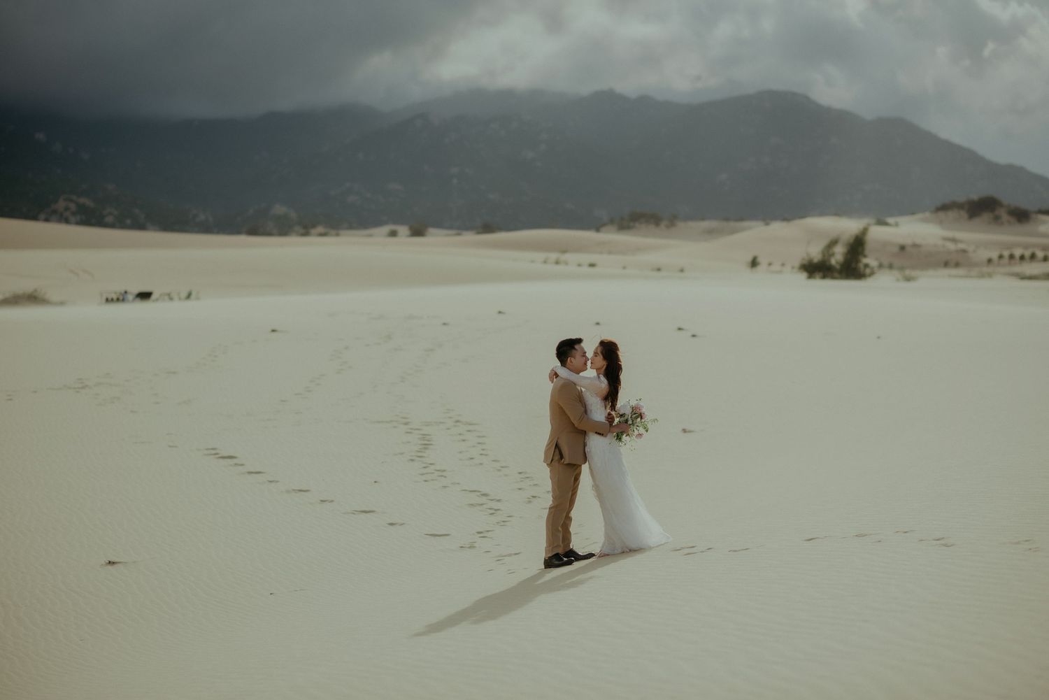 A couple shares a romantic moment on a scenic desert sand dune with misty mountains in the background.