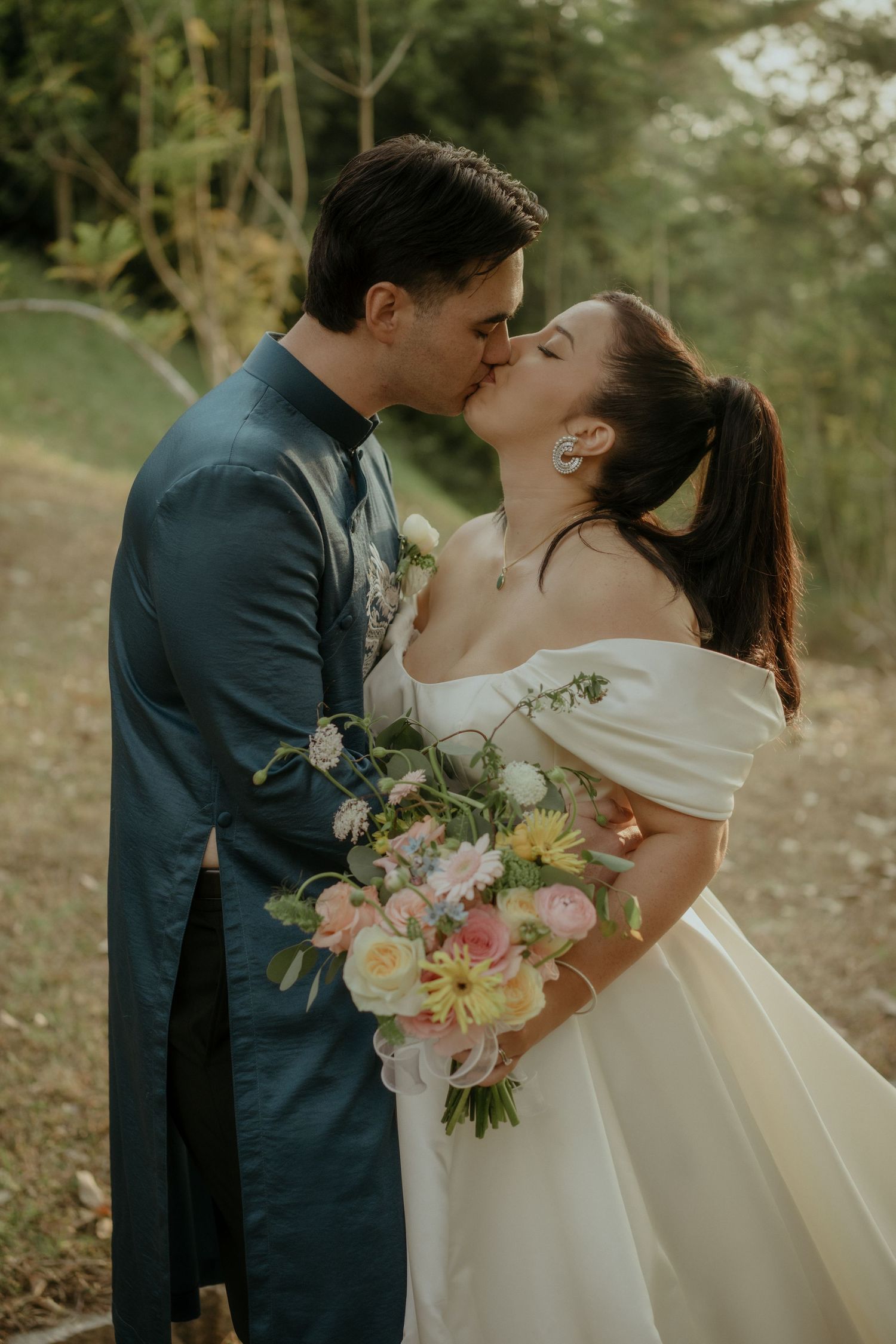 A romantic wedding couple sharing a kiss outdoors while holding a pastel bouquet of flowers on their special day.