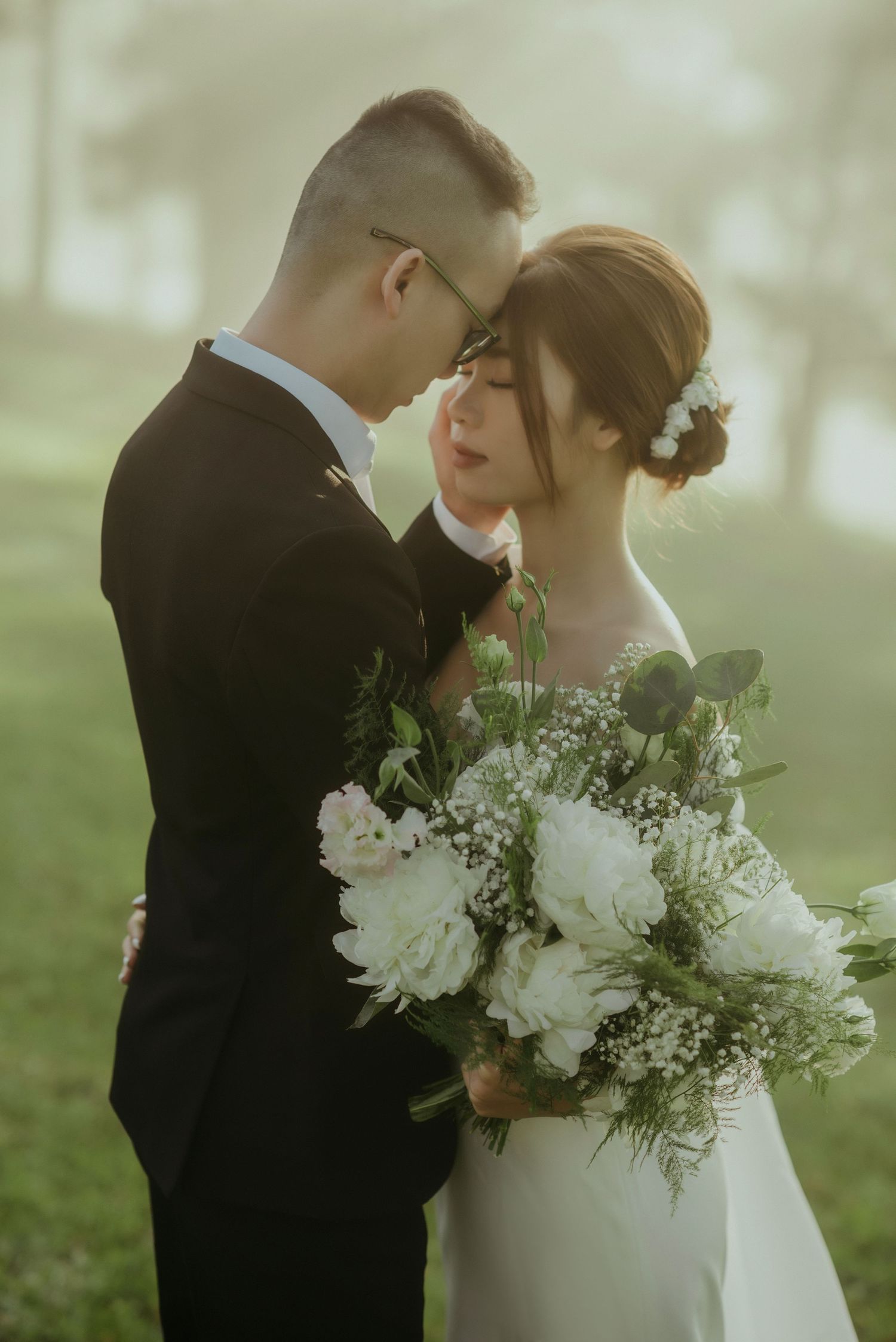 Intimate wedding couple embraces in a misty garden setting with a lush white floral bouquet and elegant attire.