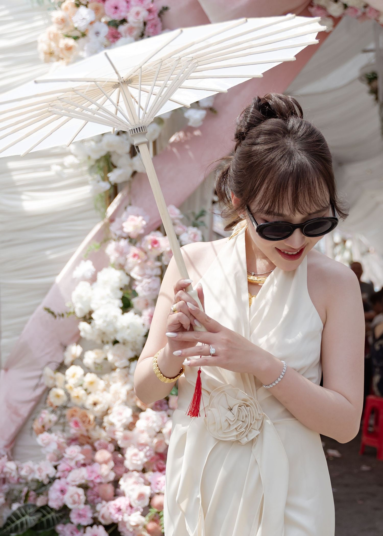 Elegant figure in white dress holding decorative paper parasol against backdrop of pink and white floral arrangements.
