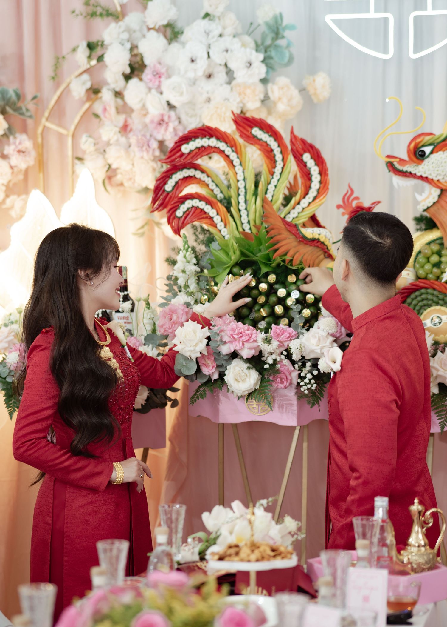 Two people in matching red outfits interact at an elegant party with pink floral decorations and festive table settings.