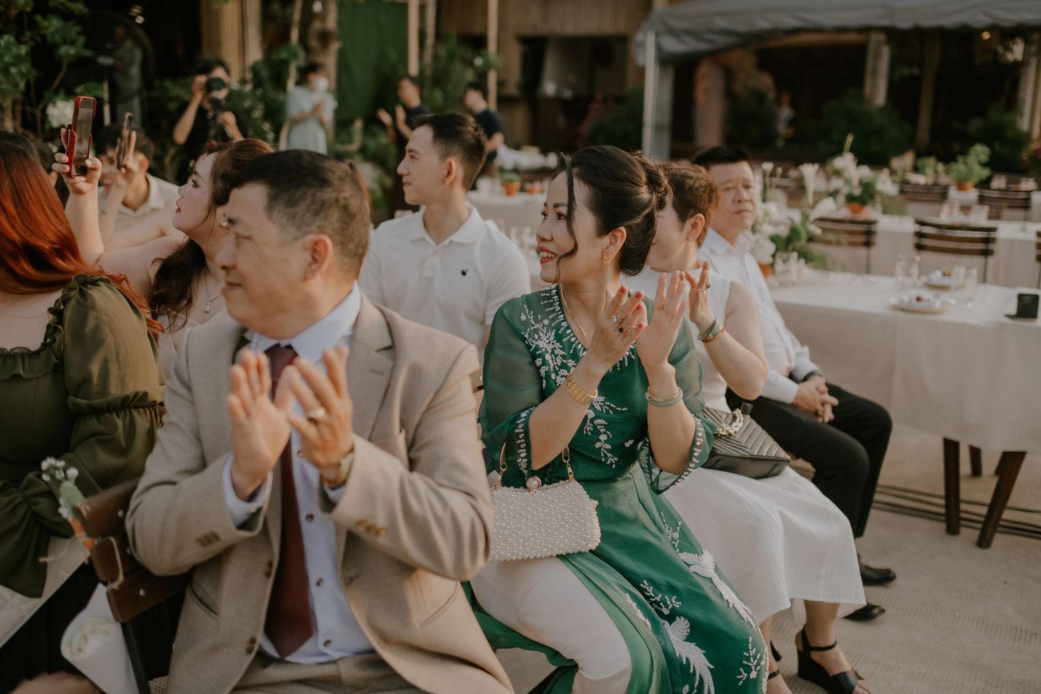 Wedding guests in formal attire applaud during an outdoor celebration.