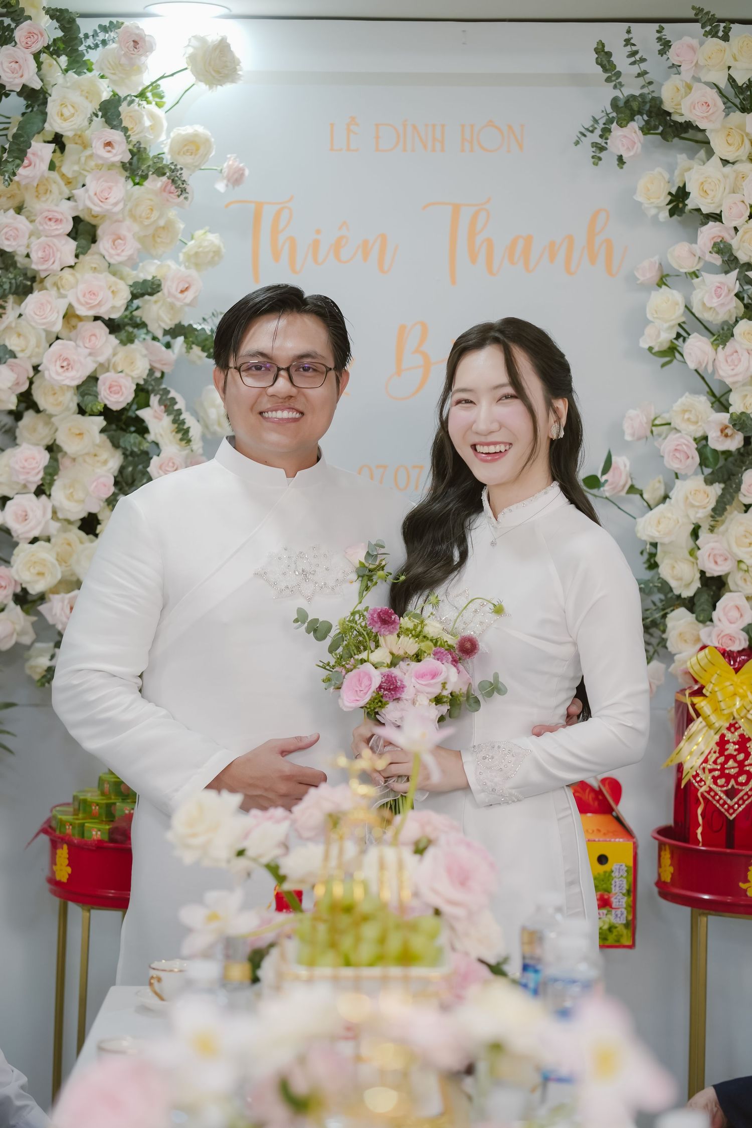 Wedding couple in white attire posing in front of floral wall with celebration decorations.